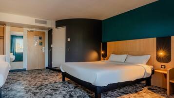 A hotel room with a bed, a desk, a chair, a door, and a wall-mounted light fixture.