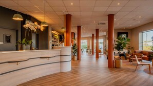 A hotel lobby with a reception desk, wooden flooring, large pillars, and indoor plants.