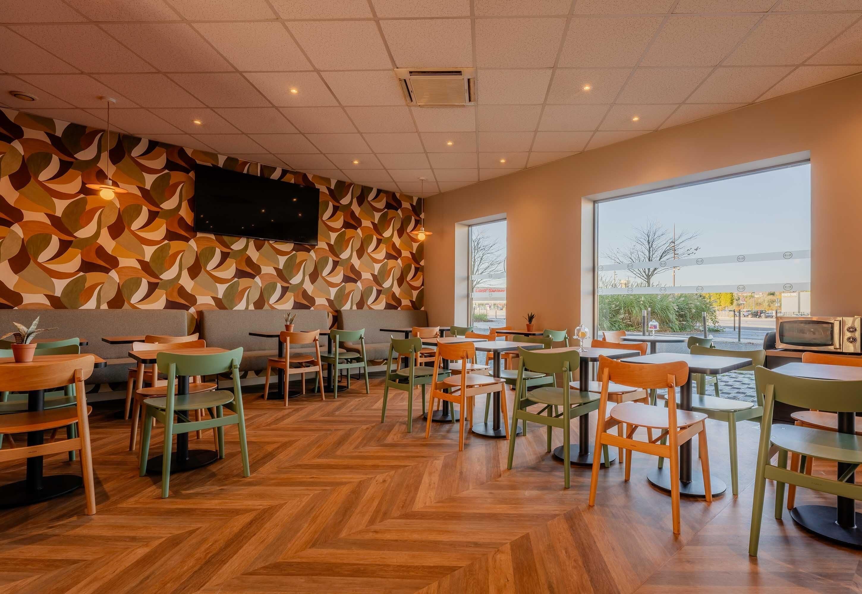 A cafe with wooden flooring, green and orange chairs, and a large window.