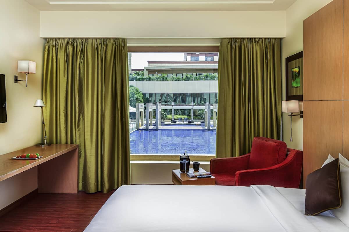 Une chambre d’hôtel avec un lit, une chaise rouge, un bureau en bois avec une lampe, une petite table avec une cafetière, et une vue sur une piscine et un immeuble.