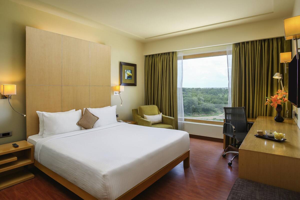 Une chambre d’hôtel avec un grand lit, un bureau agrémenté d’un vase de fleurs, une chaise, une lampe de bureau, un tableau au mur et une fenêtre avec des rideaux.