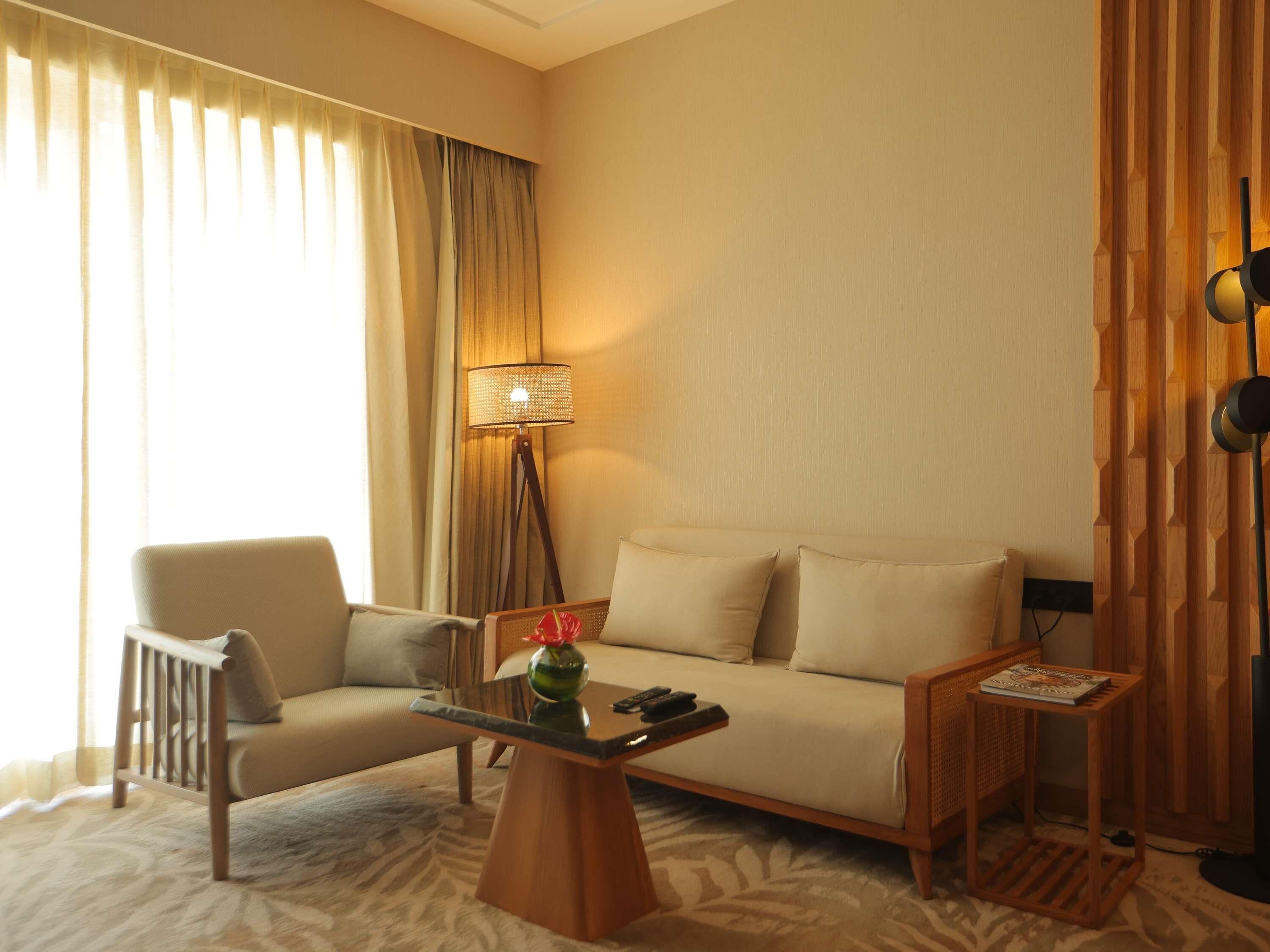 A hotel room with a sofa, armchair, and a small table with a vase and remote control.