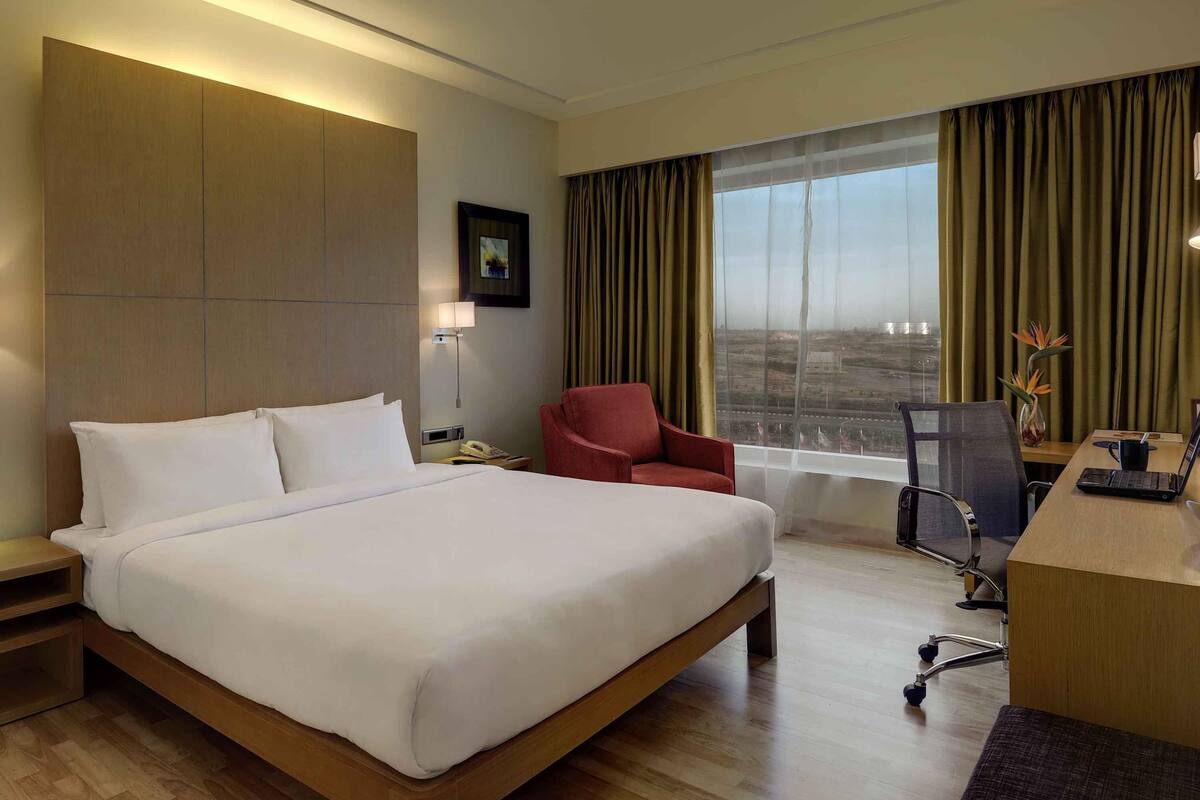 Une chambre d’hôtel avec un grand lit, un bureau avec un ordinateur portable, une chaise, une table de chevet, une lampe et une fenêtre avec des rideaux.