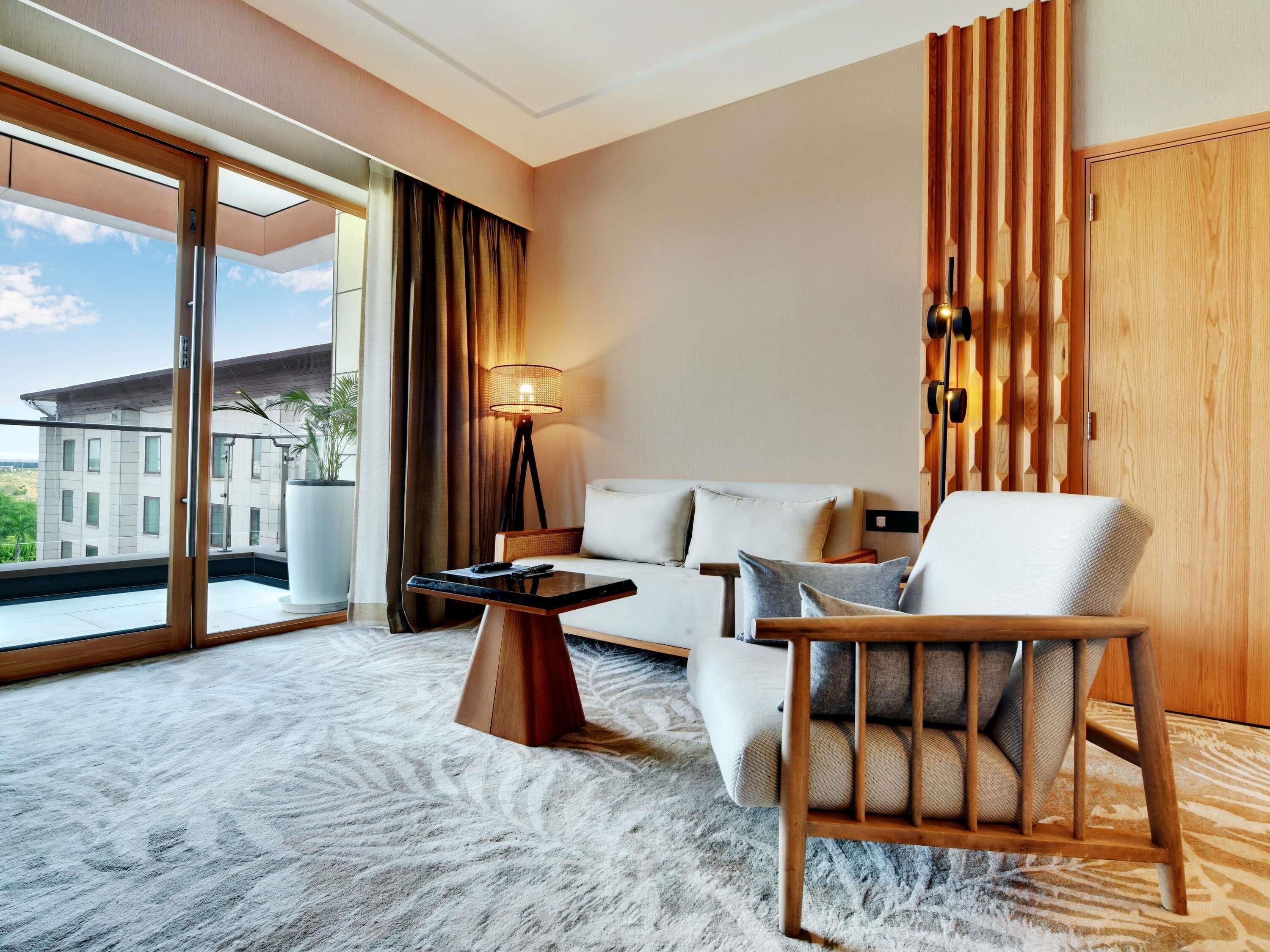Habitación de hotel con un gran ventanal corredizo, un sofá, una silla de madera, una mesita y una lámpara de pie.