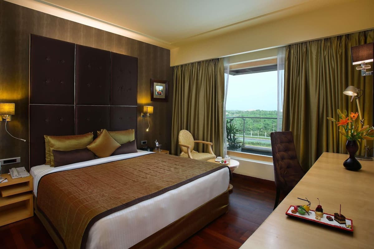 Une chambre d’hôtel avec un grand lit, un bureau avec un ordinateur portable, une chaise, une fenêtre avec des rideaux et une vue sur la verdure.