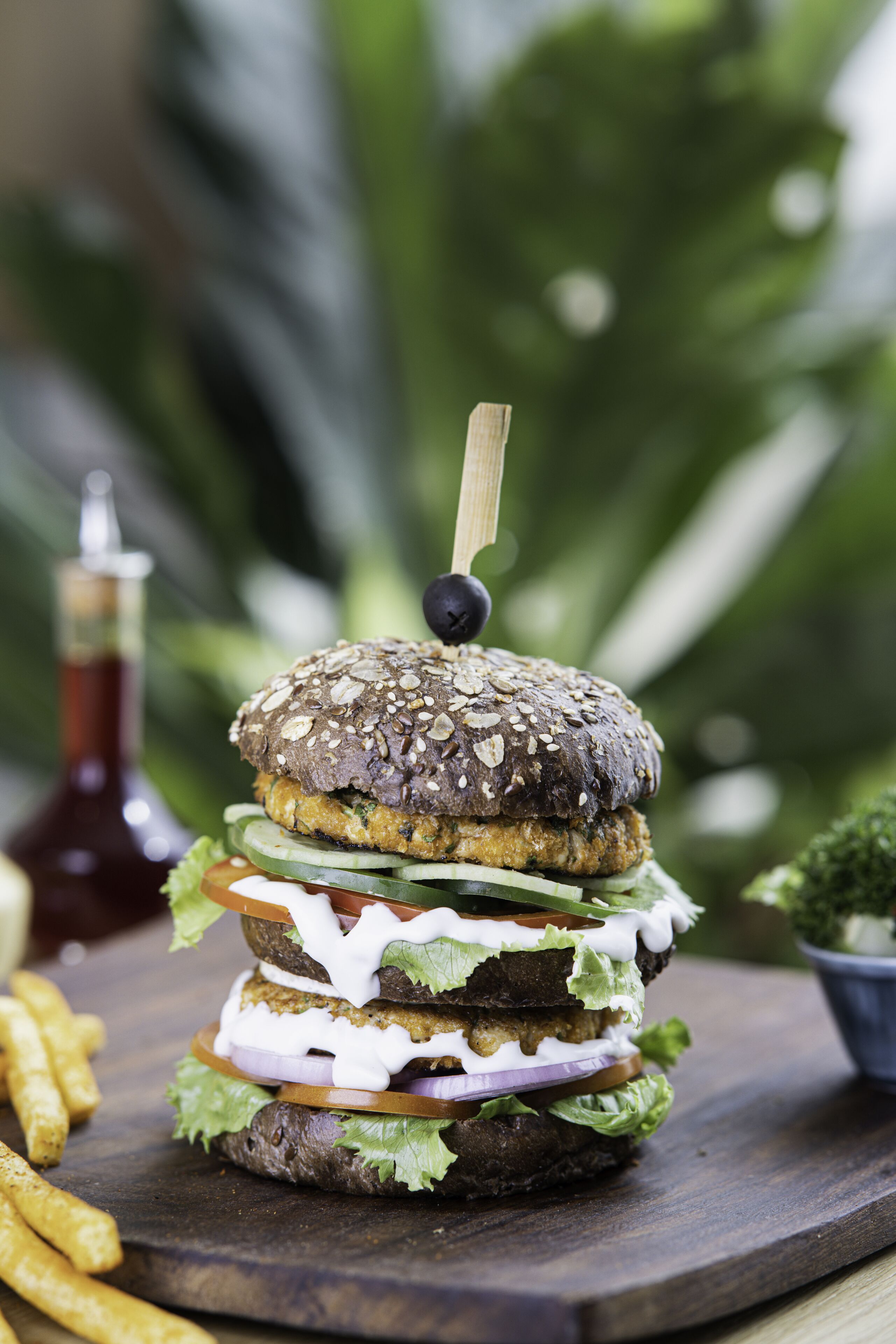 Ein mehrschichtiger Burger mit Holzspieß obenauf, umgeben von Salat-, Tomaten- und Zwiebelscheiben, mit unscharfem Grün im Hintergrund.