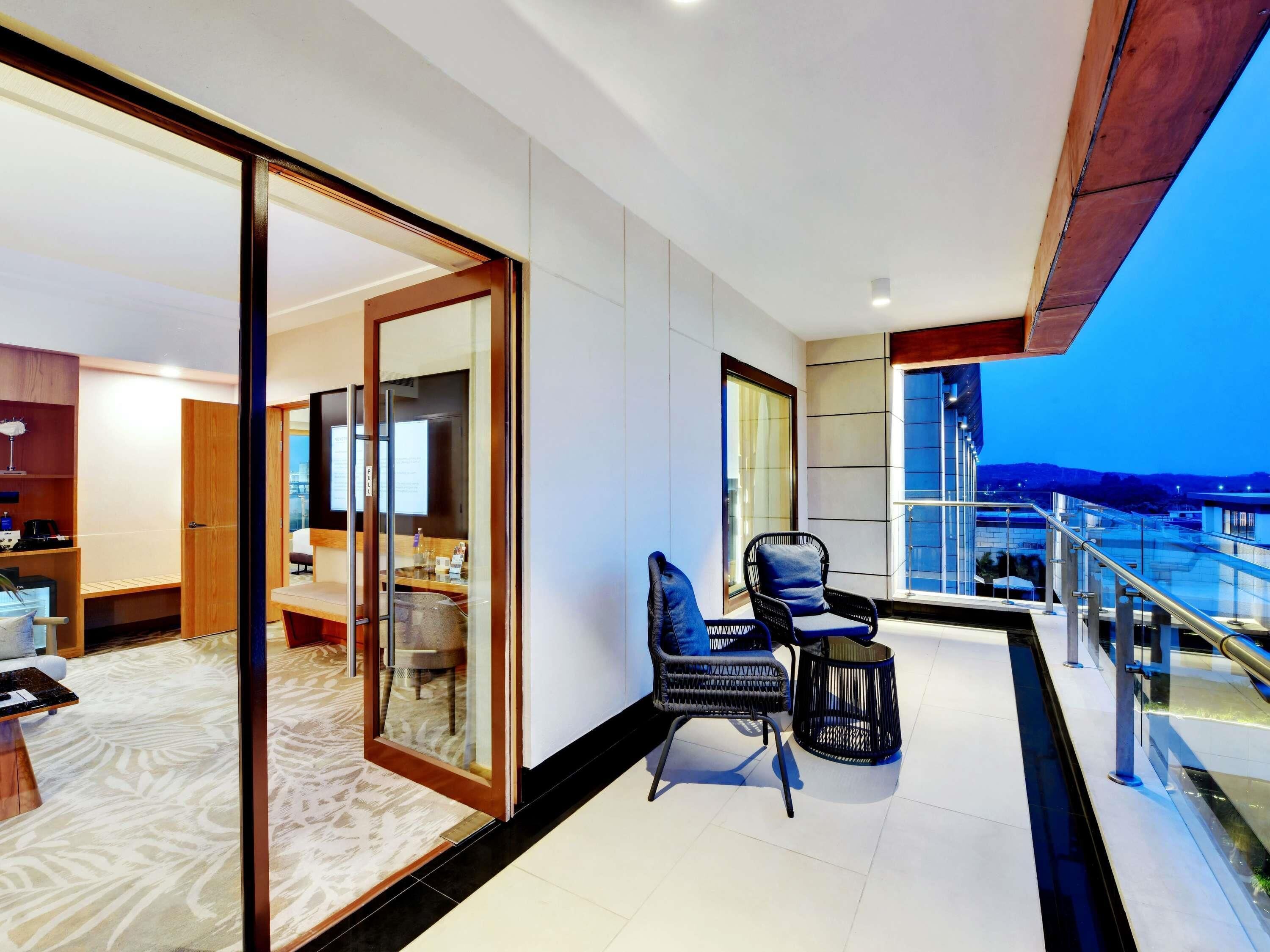 A modern hotel room with a balcony, a seating area with two chairs, and a large sliding glass door leading to a bedroom.