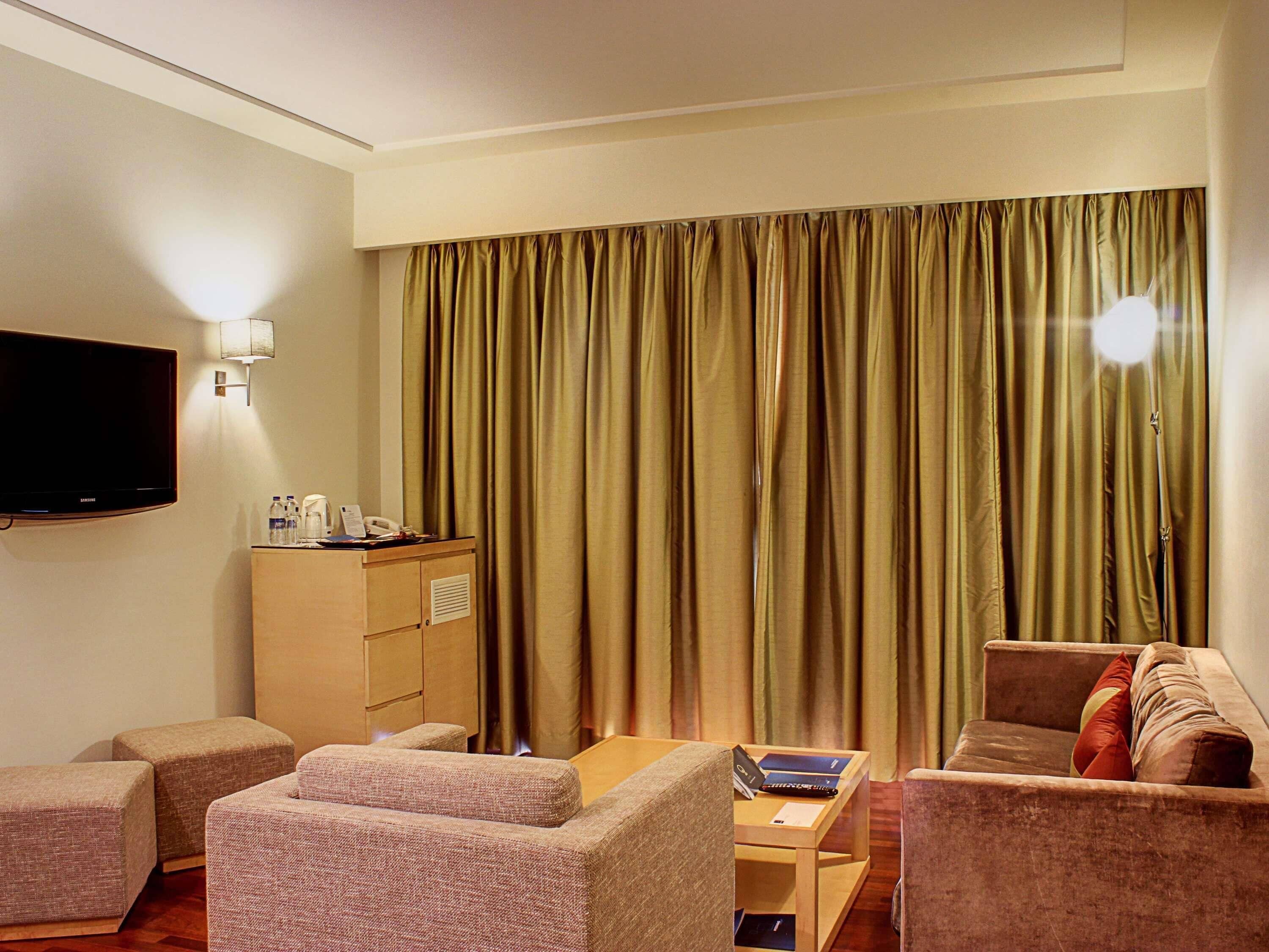 Une chambre d’hôtel équipée d’un téléviseur à écran plat, d’un mini-bar, de deux canapés, d’une petite table et d’une grande fenêtre avec des rideaux.