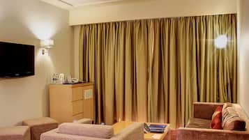 Habitación de hotel con televisor de pantalla plana, minibar, dos sofás, una mesita y un ventanal grande con cortinas.