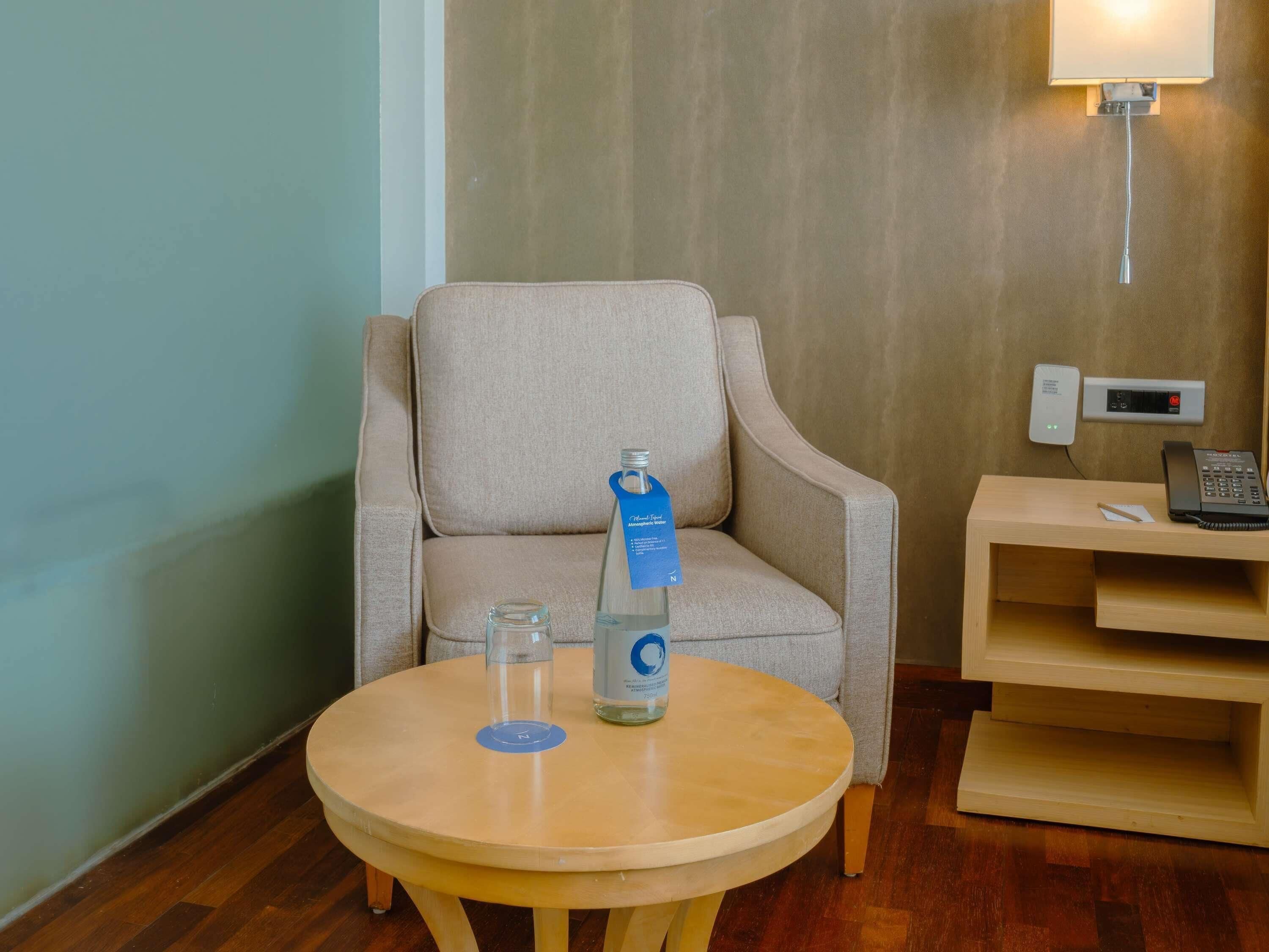 A hotel room with a round wooden table, a beige armchair, a bottle of water, a glass, a telephone, and a wall-mounted control panel.