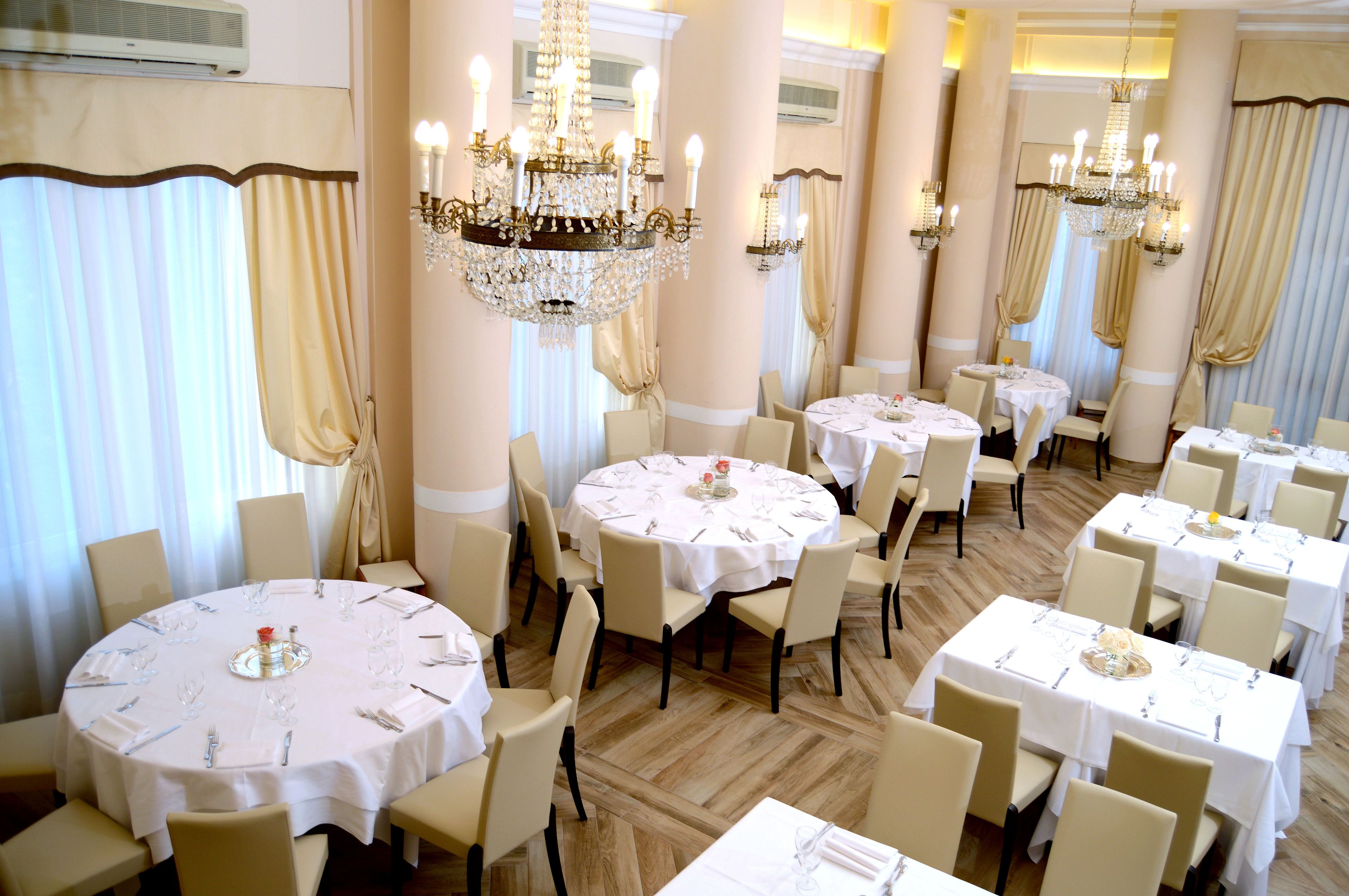 A formal dining room with multiple round tables, each set with white tablecloths, chairs, and chandeliers.