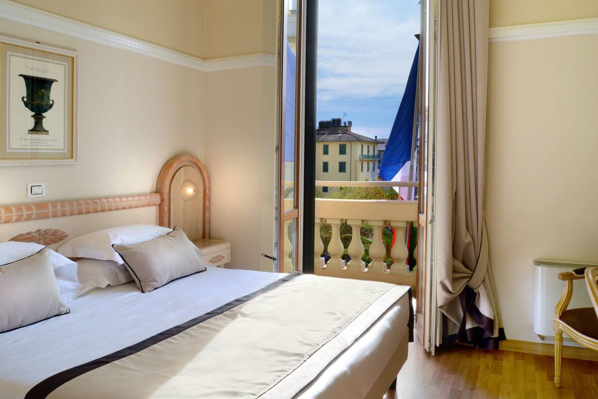 Ein Hotelzimmer mit Bett, Stuhl, einem Fenster mit Vorhängen und einem Balkon.