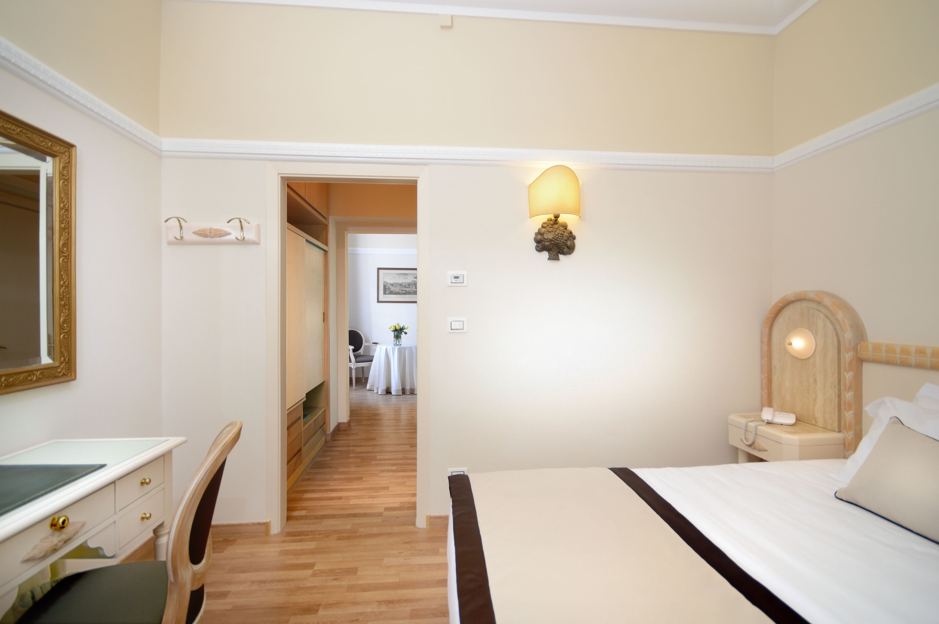A hotel room with a bed, a desk, a chair, a mirror, and a hallway leading to another room.