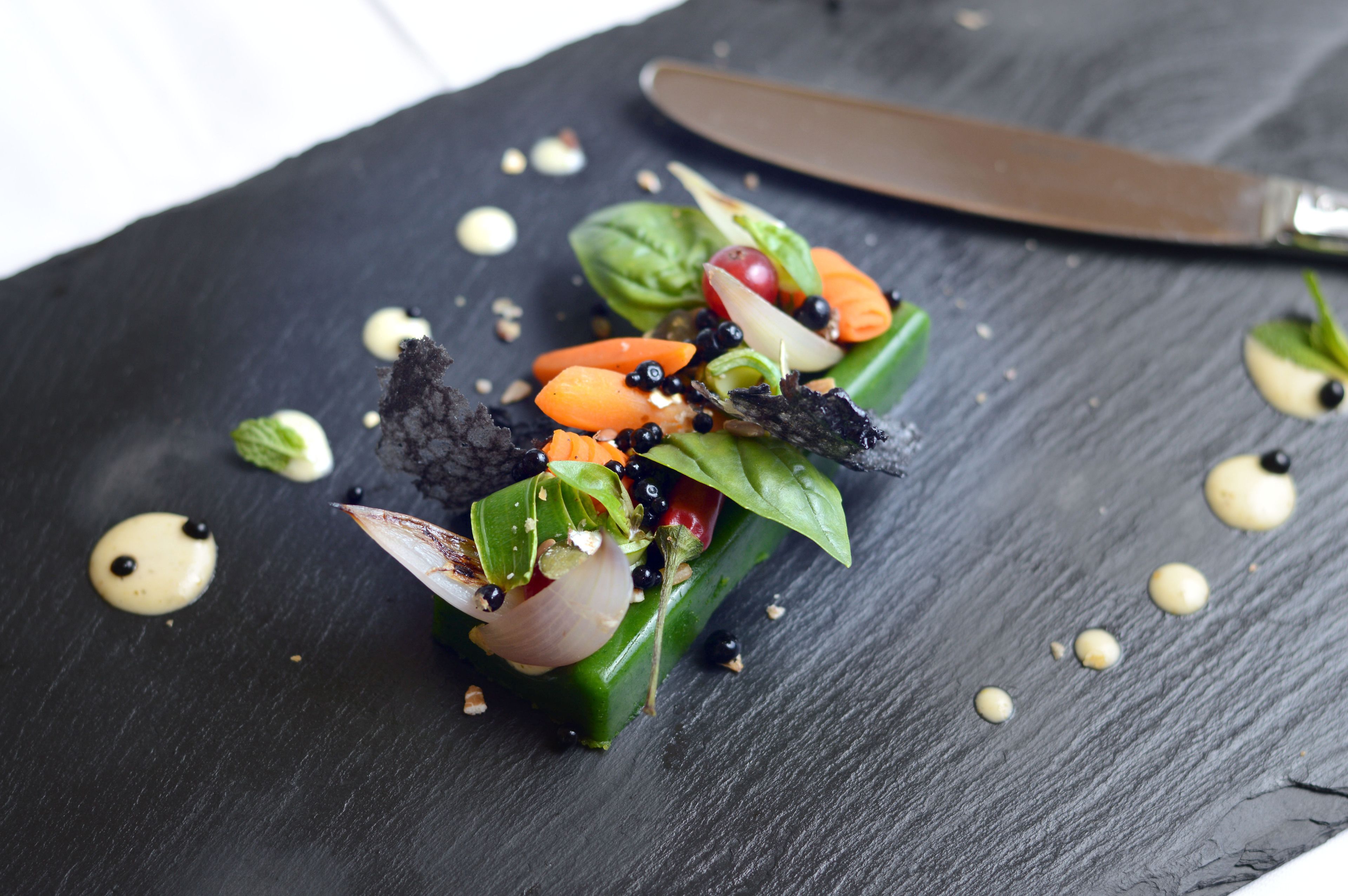 A black slate serving board with a green vegetable dish, black olives, and a knife.