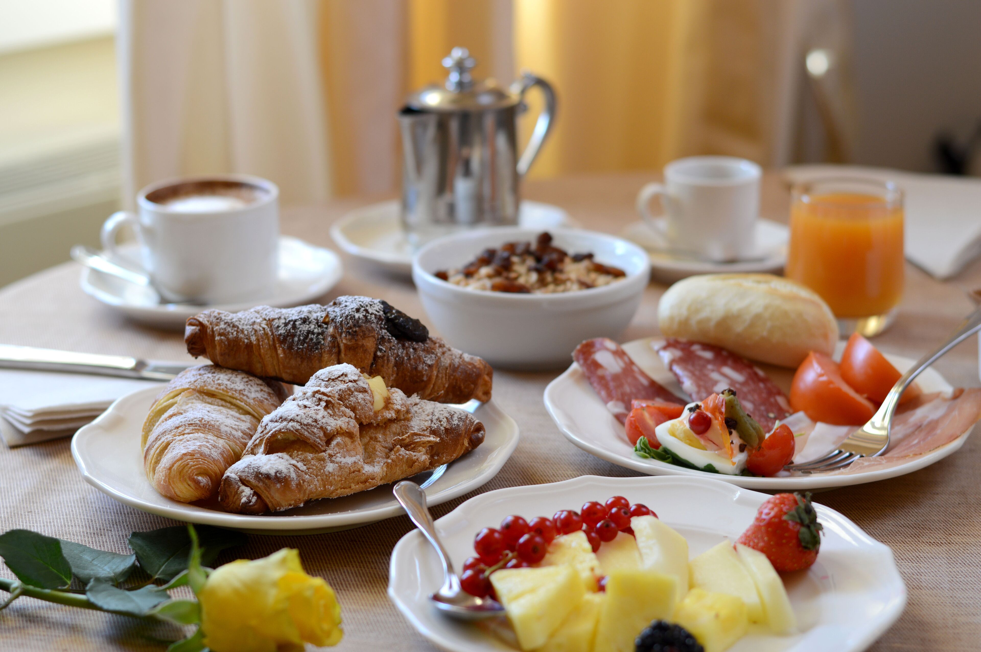 Une table de petit-déjeuner avec des croissants, des fruits, une tasse de café, un verre de jus de fruits et une théière en argent.