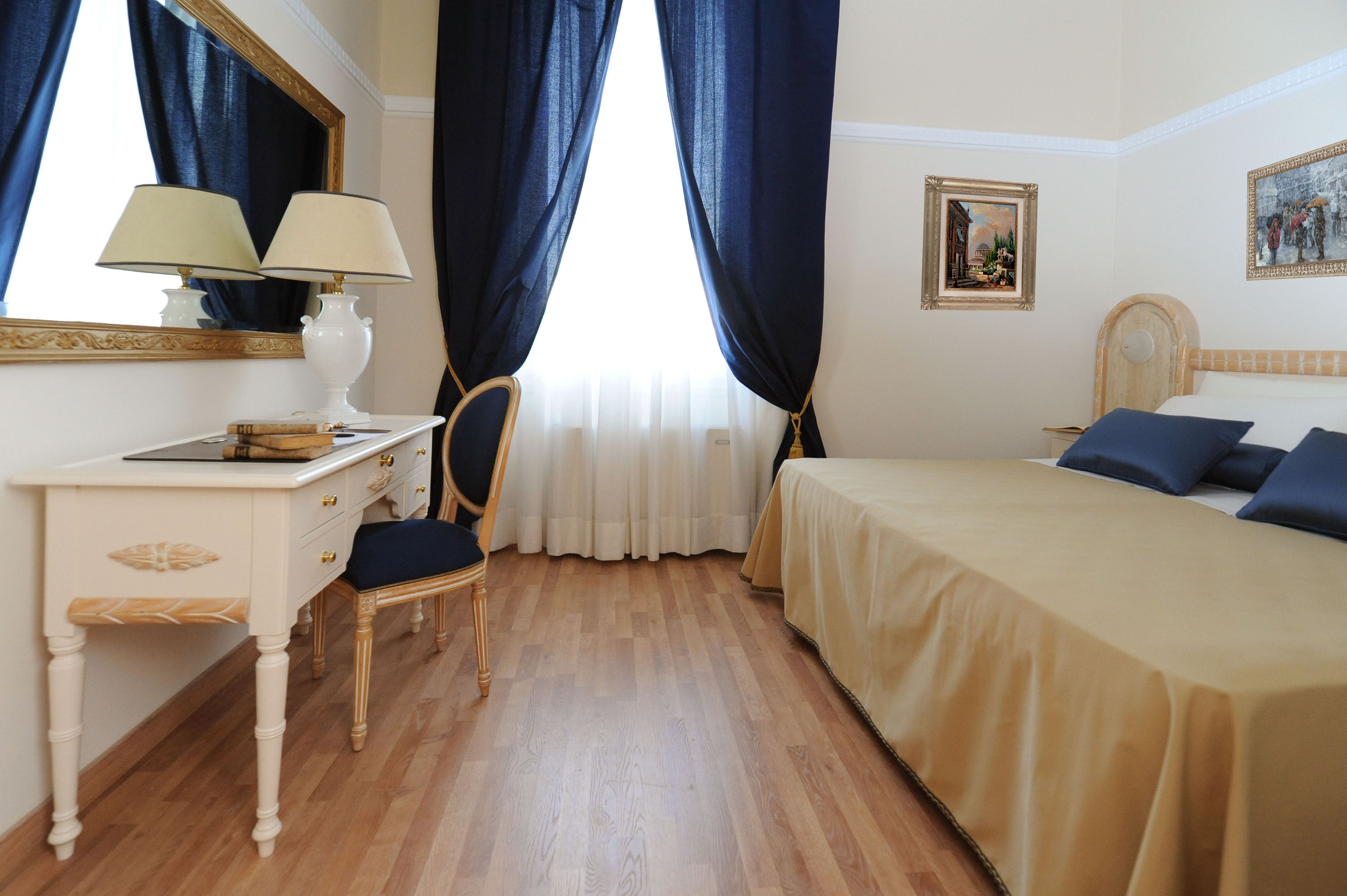 Habitación de hotel con suelo de madera, un escritorio con lámpara, una silla, una cama con cabecero y dos cuadros en la pared.