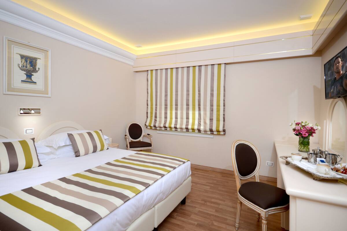 Ein Hotelzimmer mit einem Bett, einem Schreibtisch, einem Stuhl, einem Fenster mit gestreifter Vorhang und einer Blumenvase.