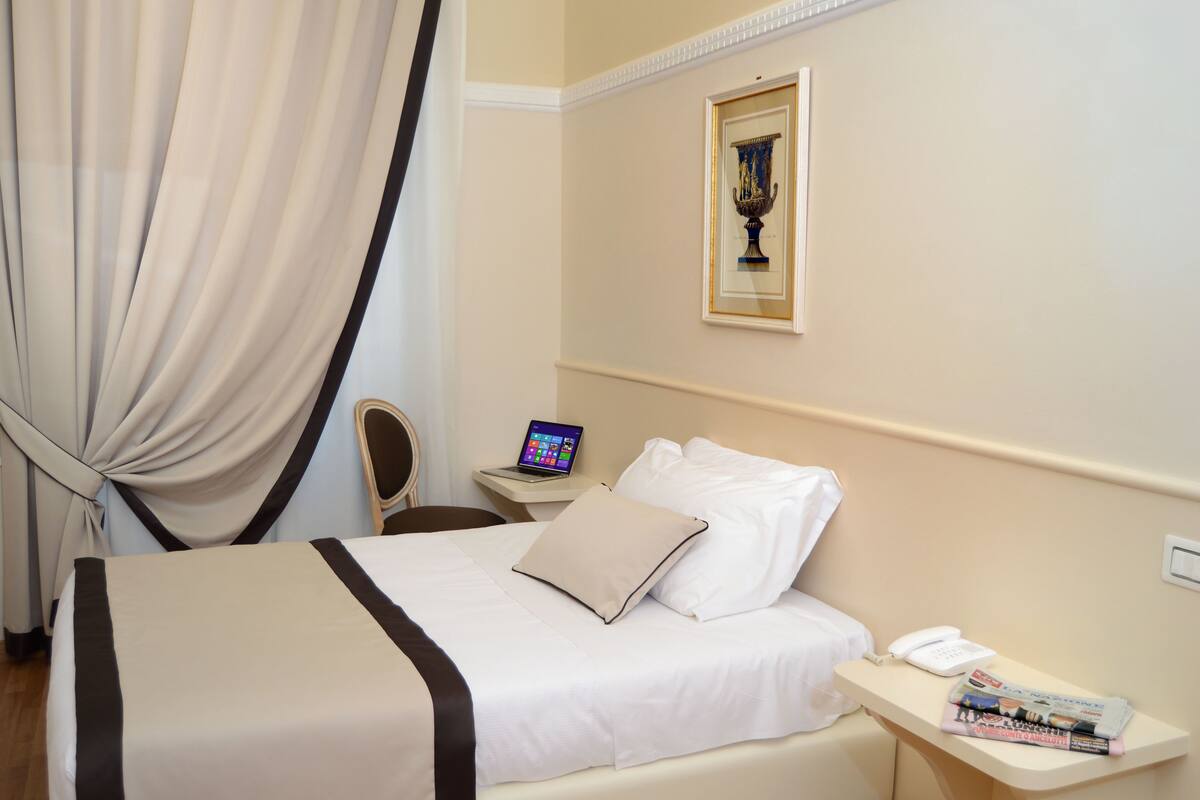 Ein Hotelzimmer mit einem Bett, einem Schreibtisch mit Laptop, einem Telefon und einer Zeitschrift auf dem Nachttisch.