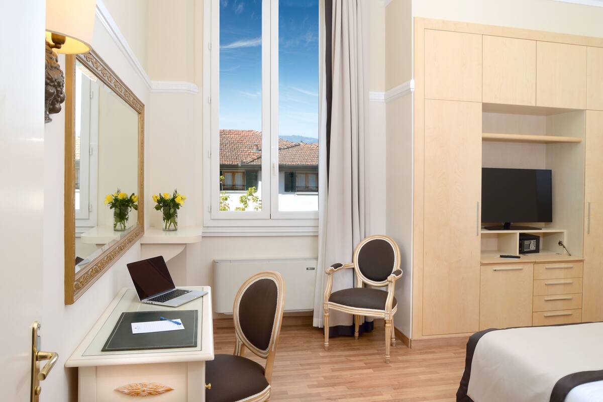 Ein Hotelzimmer mit Schreibtisch, Laptop, Stuhl, Fernseher und einem großen Fenster mit Blick auf Gebäude.