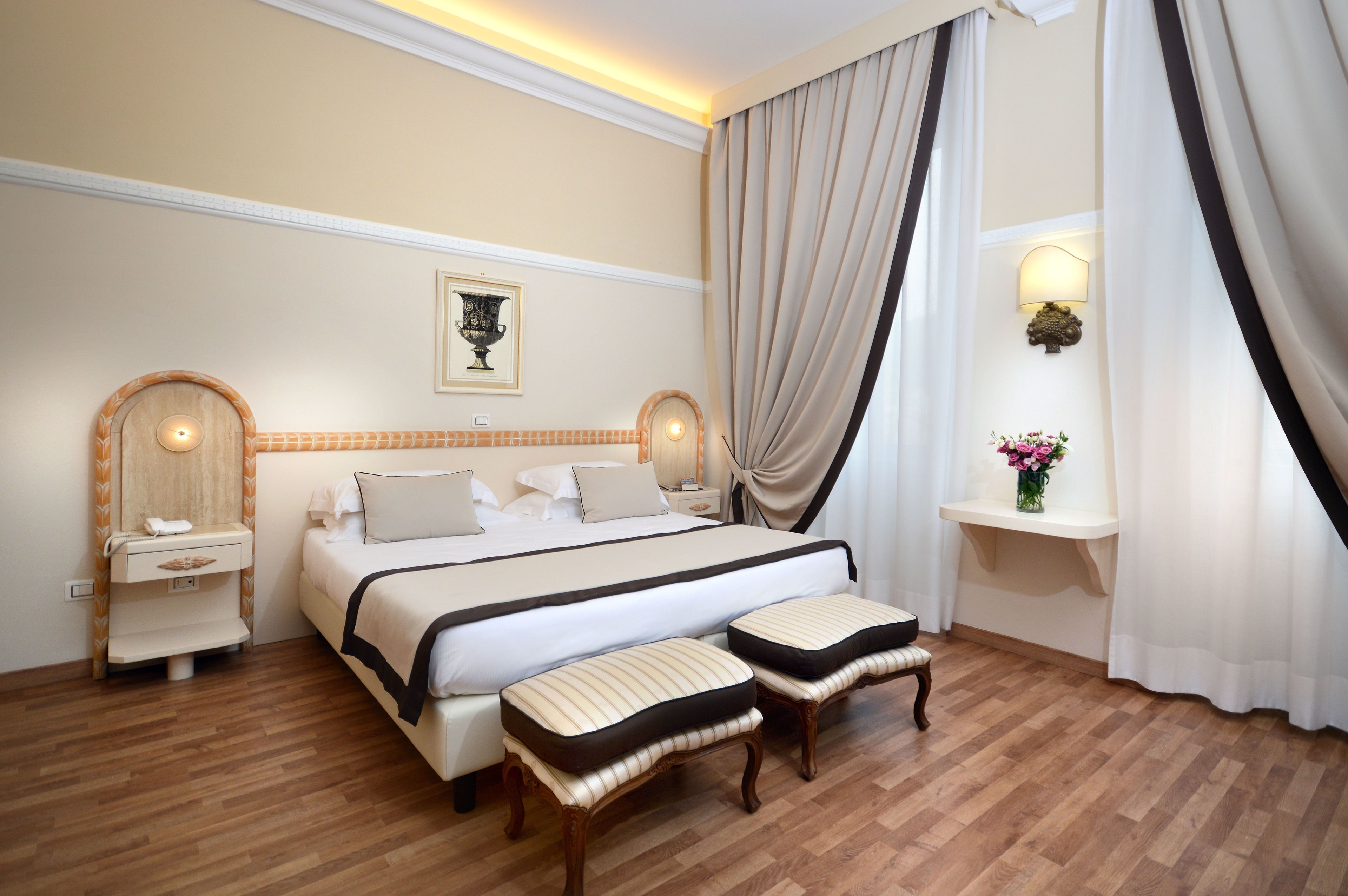 Een hotelkamer met een groot bed, twee nachtkastjes, een wandlamp, een plank met een vaas en een raam met gordijnen.