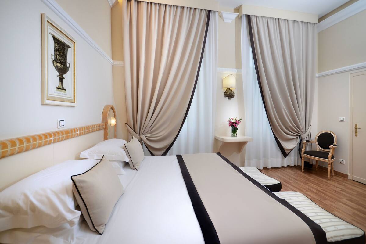 Ein Hotelzimmer mit einem großen Bett, zwei Nachttischen, einem Sessel und Vorhängen.