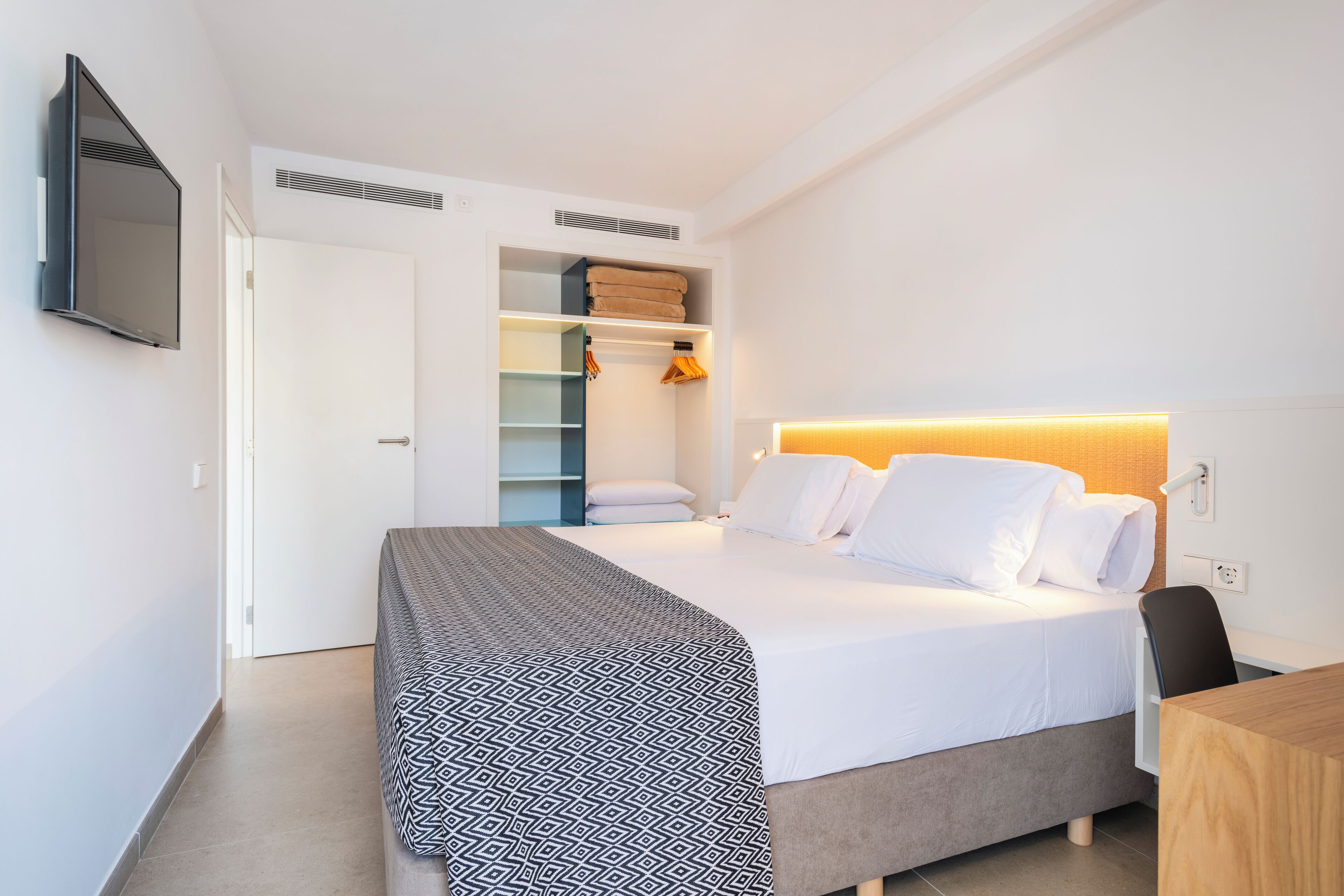 A modern hotel room with a neatly made bed, a built-in wardrobe, and a flat-screen TV mounted on the wall.