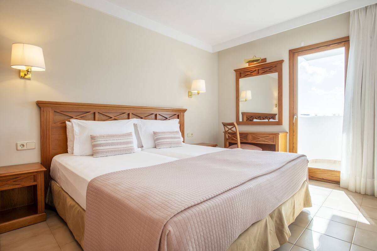 A hotel room with a large bed, bedside tables, a desk, a chair, a mirror, and a view of the sea.