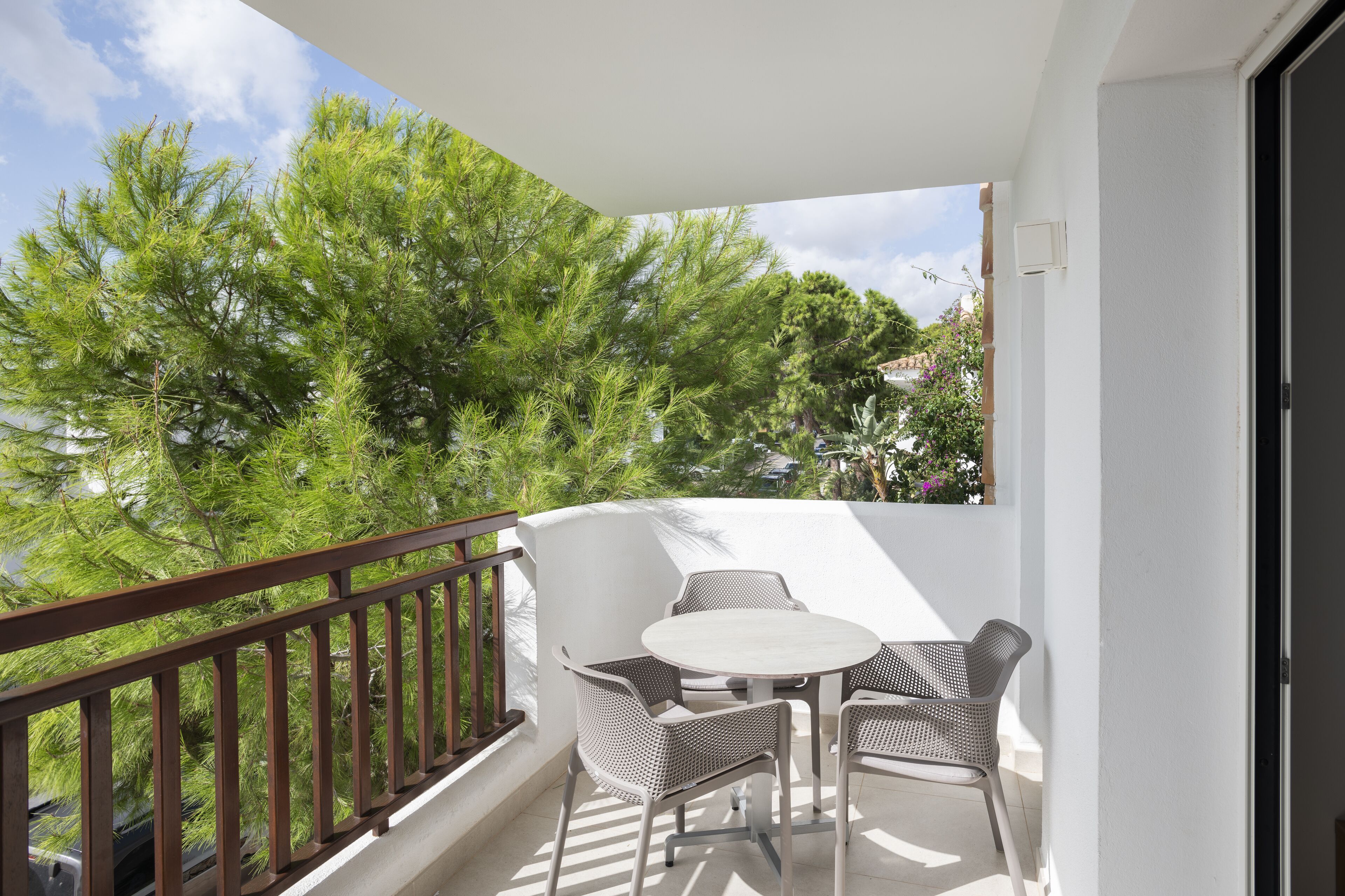 A balcony with a white table and wicker chairs, overlooking a garden with trees and flowers.