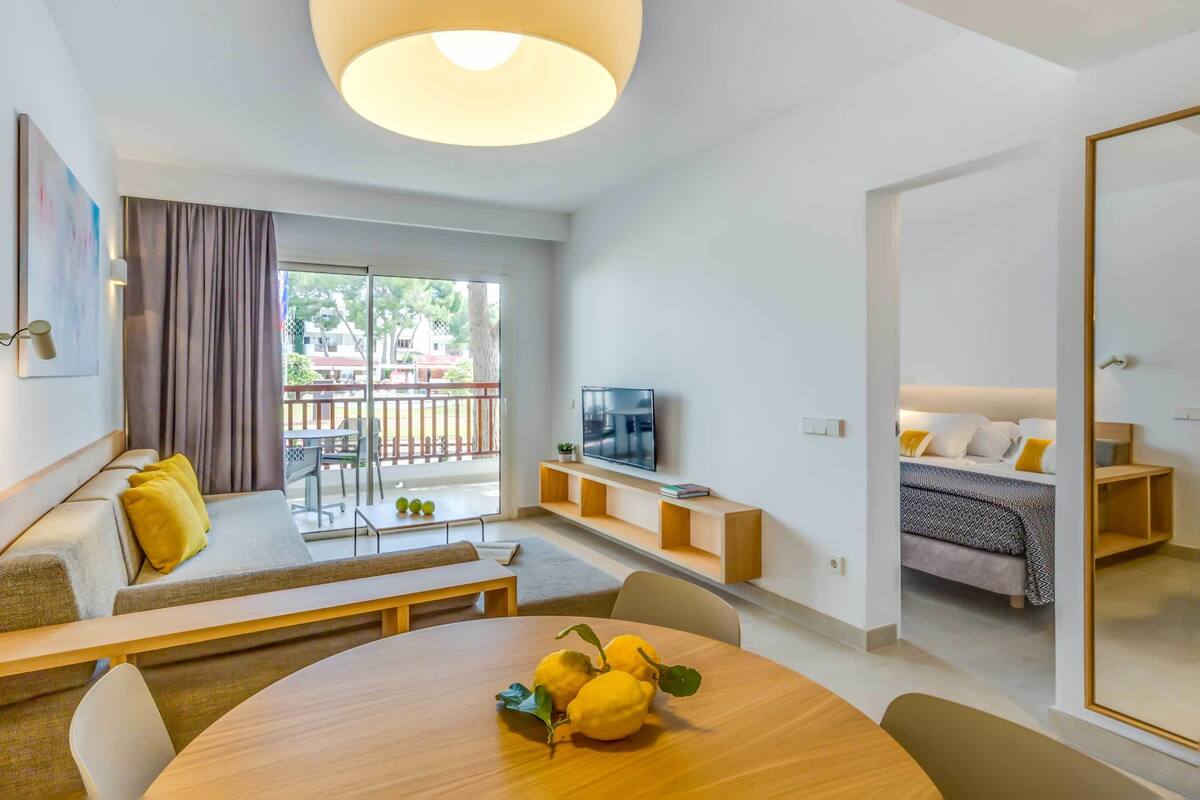 A modern living room with a round wooden table, a sofa with yellow cushions, a TV on a wooden shelf, and a balcony with a view of the outside.
