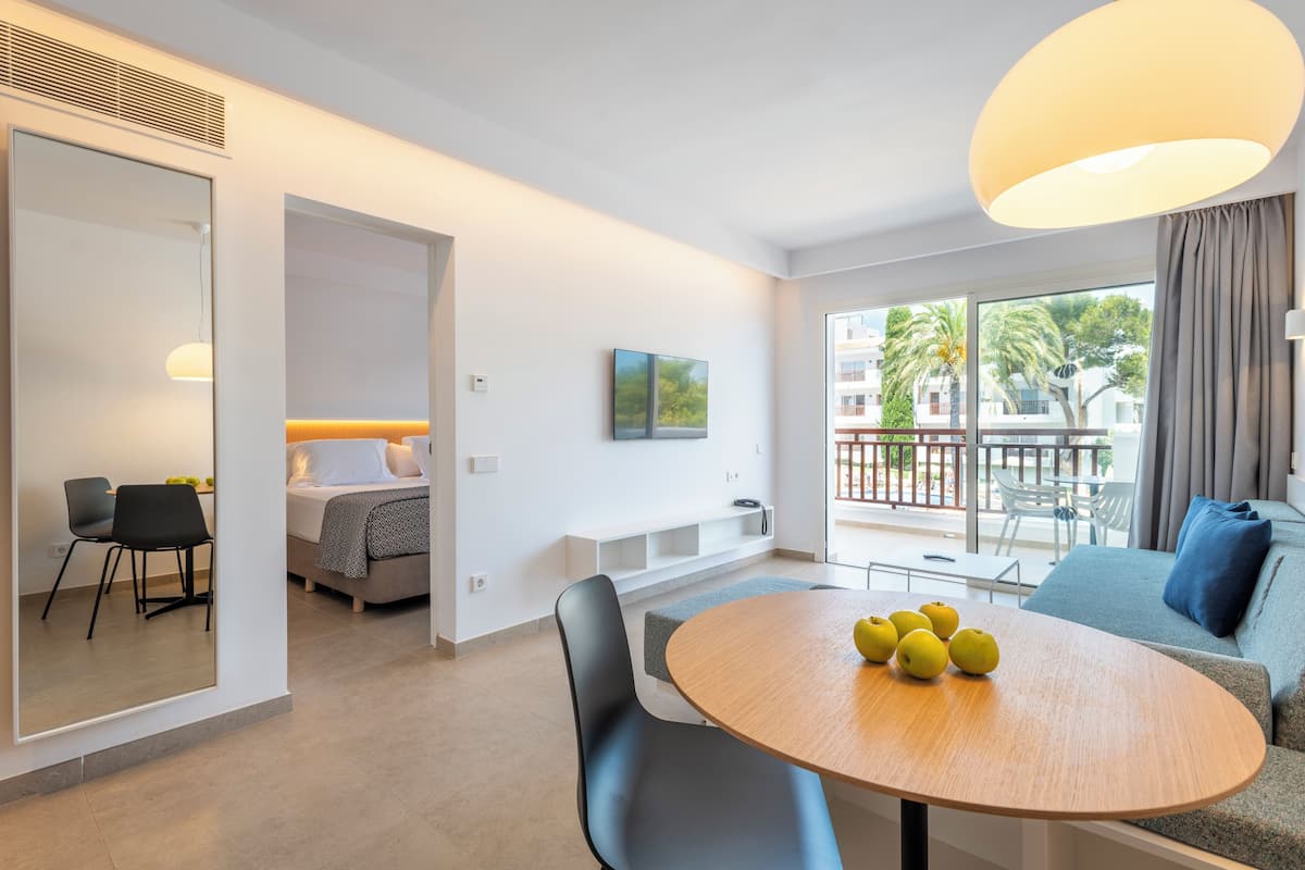 A modern living room with a round wooden table, a sofa, and a balcony with a view of palm trees and buildings.