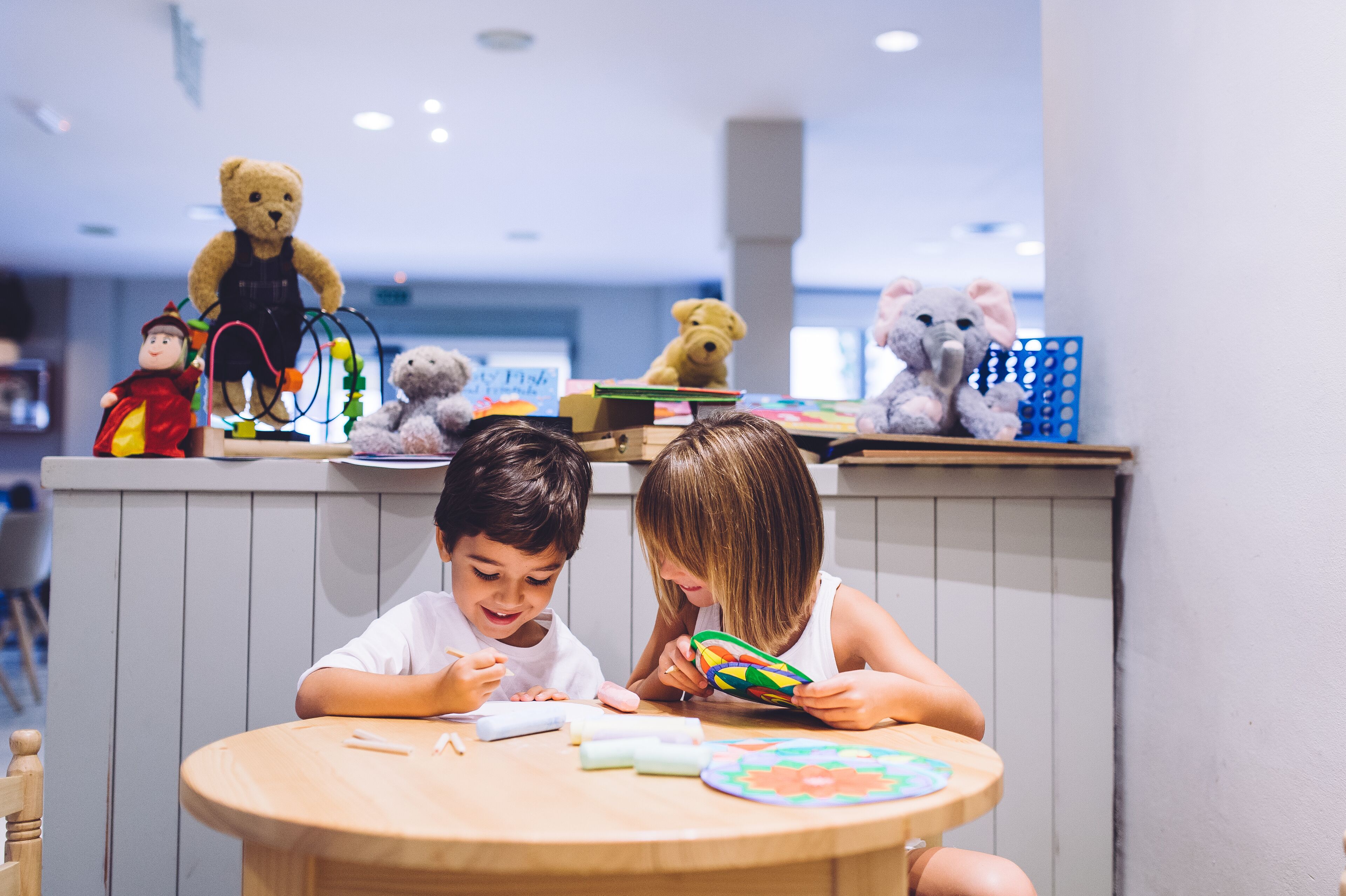 Two children at a table with toys and books, in a room with a wooden table and chairs.