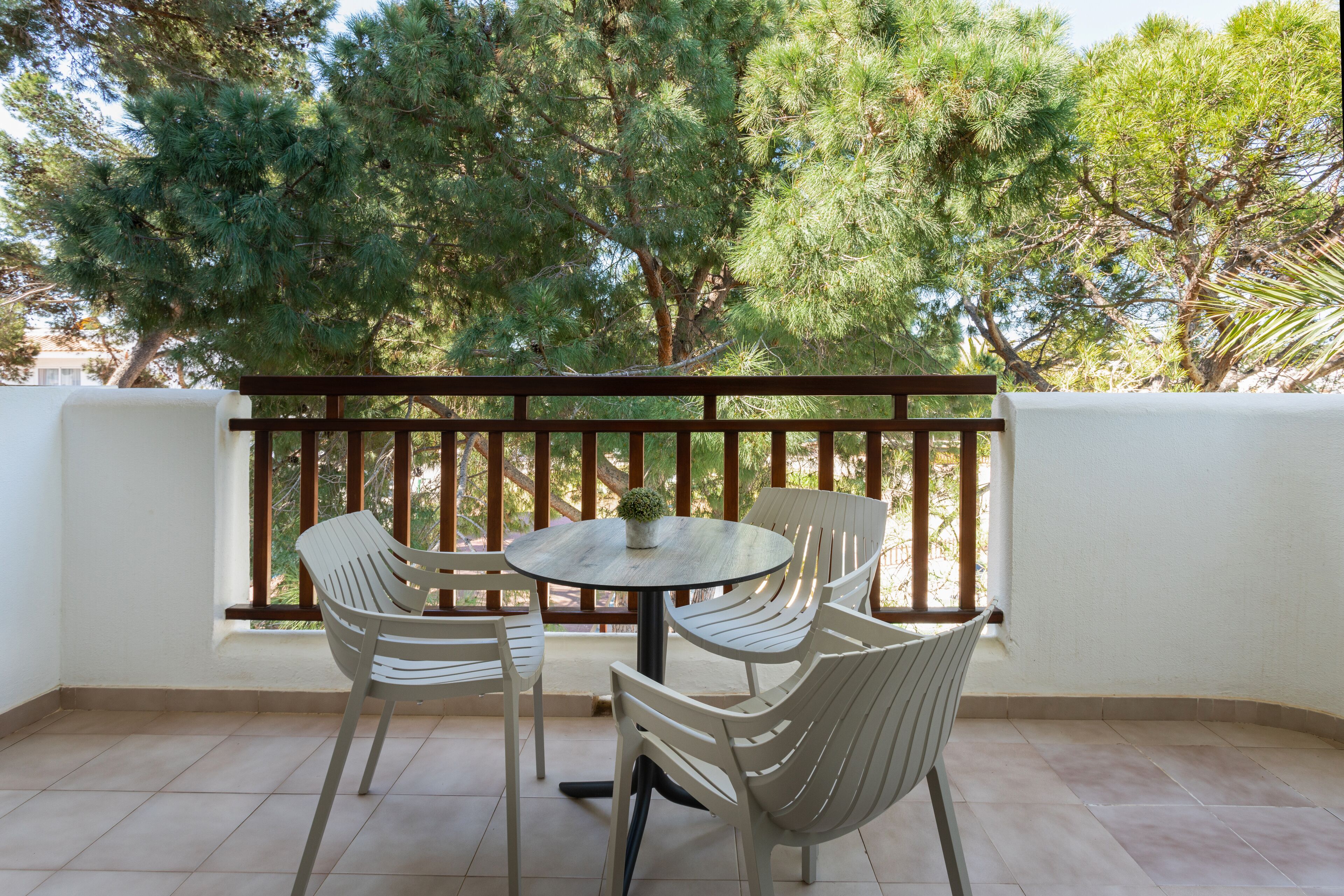 Un balcón con una mesa redonda y cuatro sillas, rodeado de baranda de madera, con vistas a un jardín con árboles.
