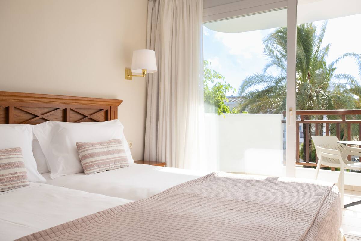 A hotel room with a large bed, a wooden headboard, a white lamp, and a view of palm trees through a balcony.