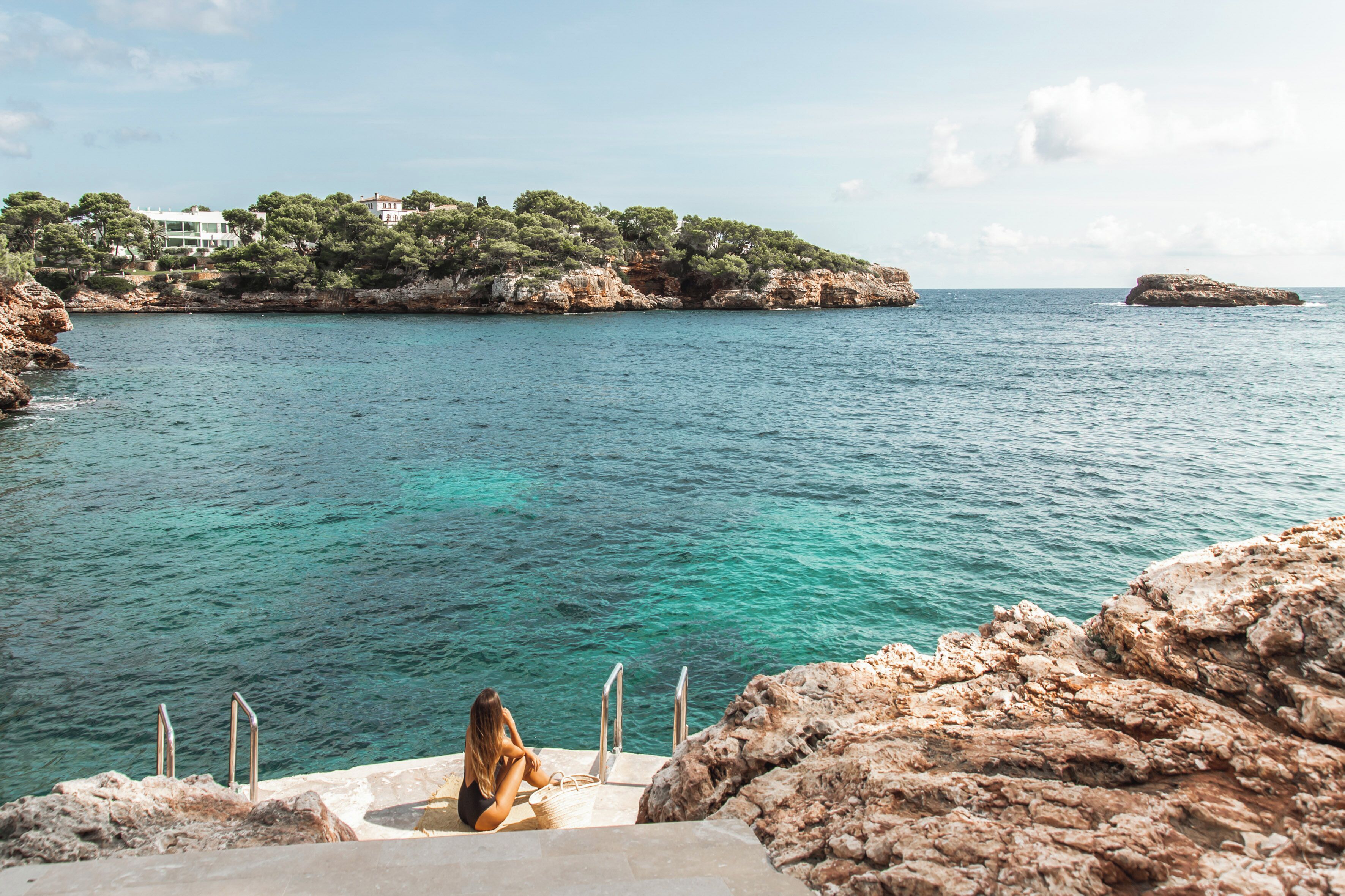 A woman sitting on a stone bench overlooking a clear blue sea with rocky cliffs and buildings in the background.