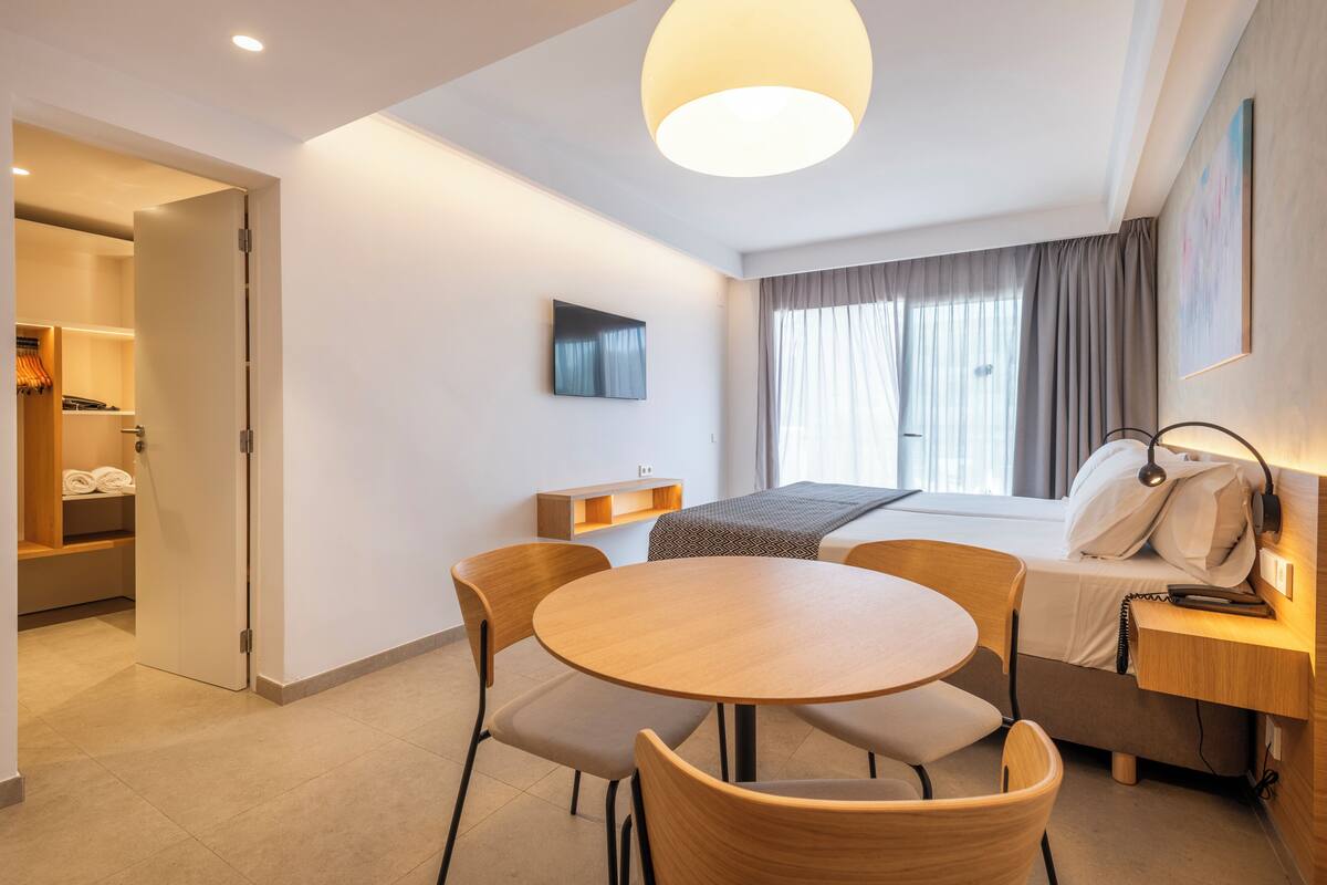 A modern hotel room with a round wooden table, two chairs, a bed with a headboard, a bedside table with a lamp, a TV mounted on the wall, and a window with curtains.