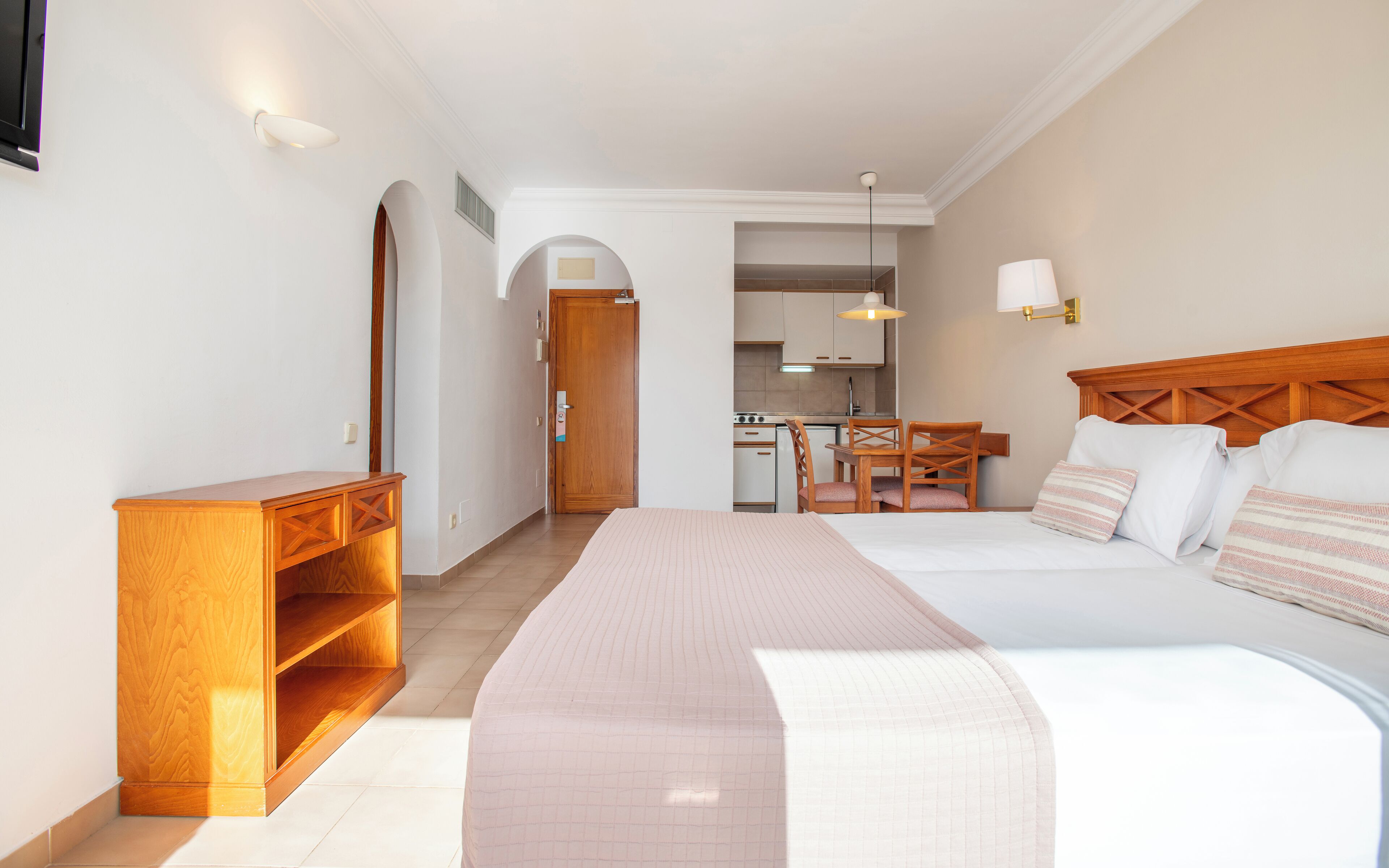 Une chambre d’hôtel avec un lit, une table de chevet en bois, un bureau, une chaise et, au fond, une petite cuisine.