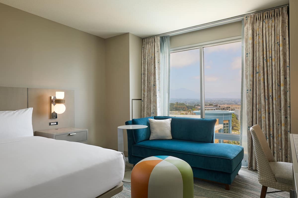 Habitación de hotel con cama, sofá azul, mesita y vista a la ciudad a través de amplias ventanas.