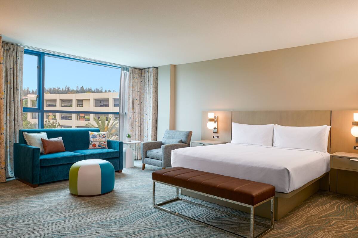 Una habitación de hotel con una cama grande, un sofá azul, un sillón gris, una mesita y una ventana con cortinas.