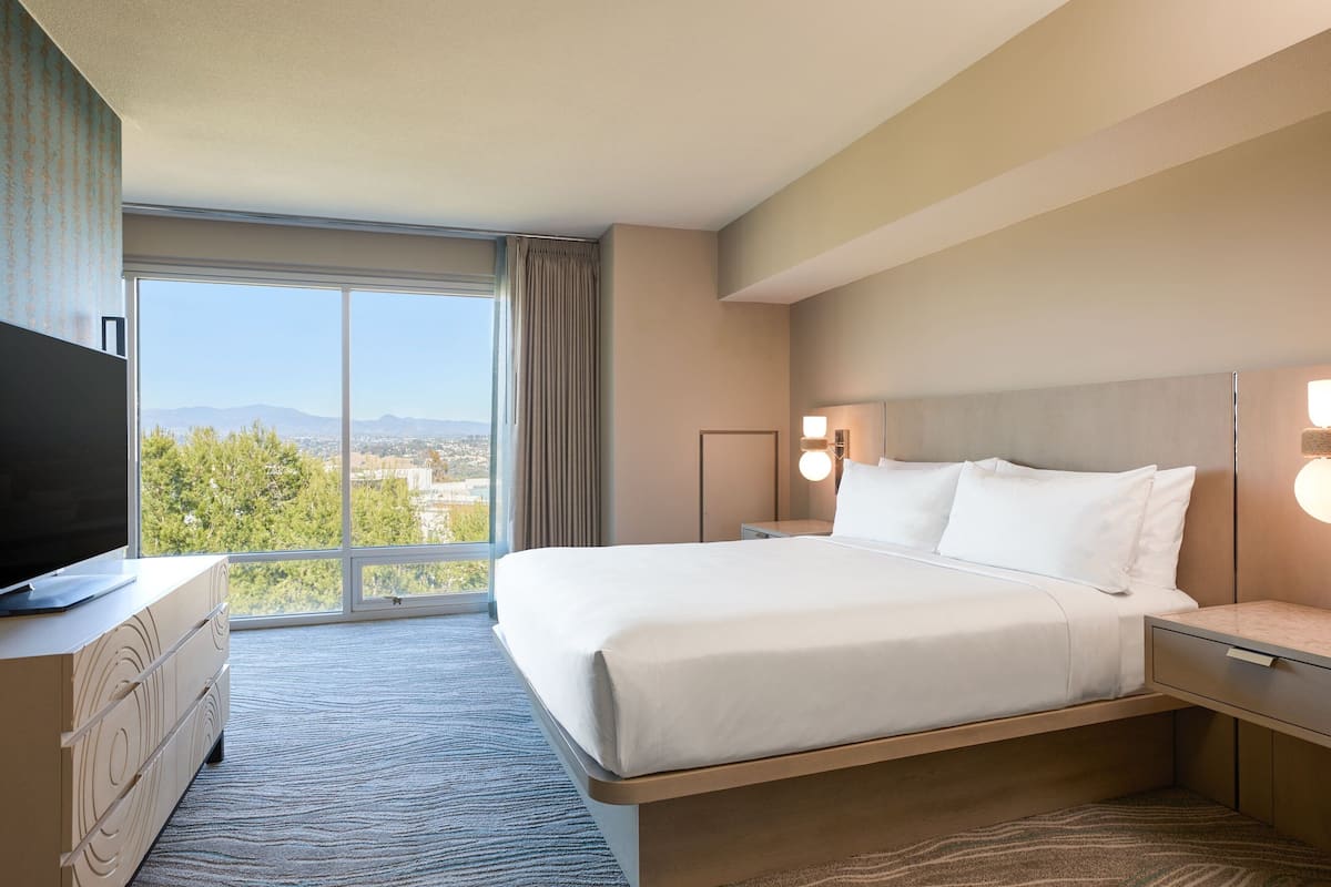 Habitación de hotel con una cama grande, un televisor sobre una cómoda, una mesita de noche y vistas de la ciudad a través de un ventanal amplio.