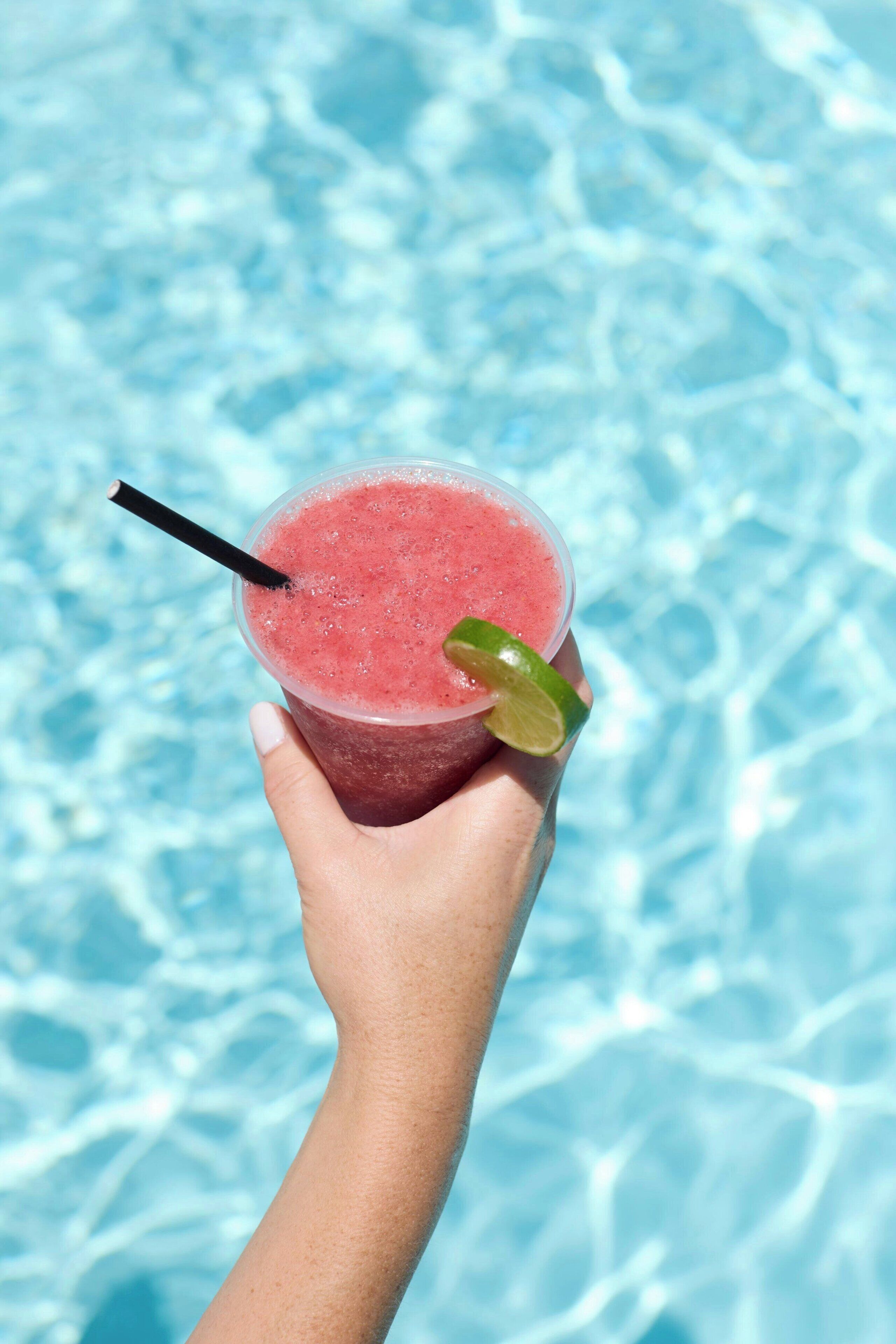 A hand holding a pink drink with a lime wedge and a straw, against a poolside background.
