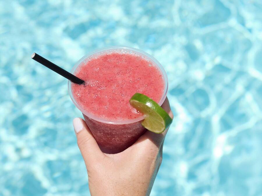 A hand holding a pink drink with a lime wedge and a straw, against a poolside background.