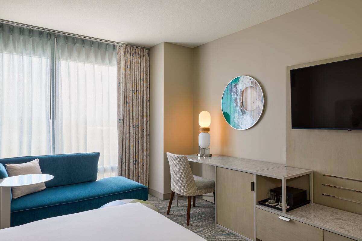 A hotel room with a bed, a blue sofa, a desk with a lamp, a TV, and a large window with curtains.