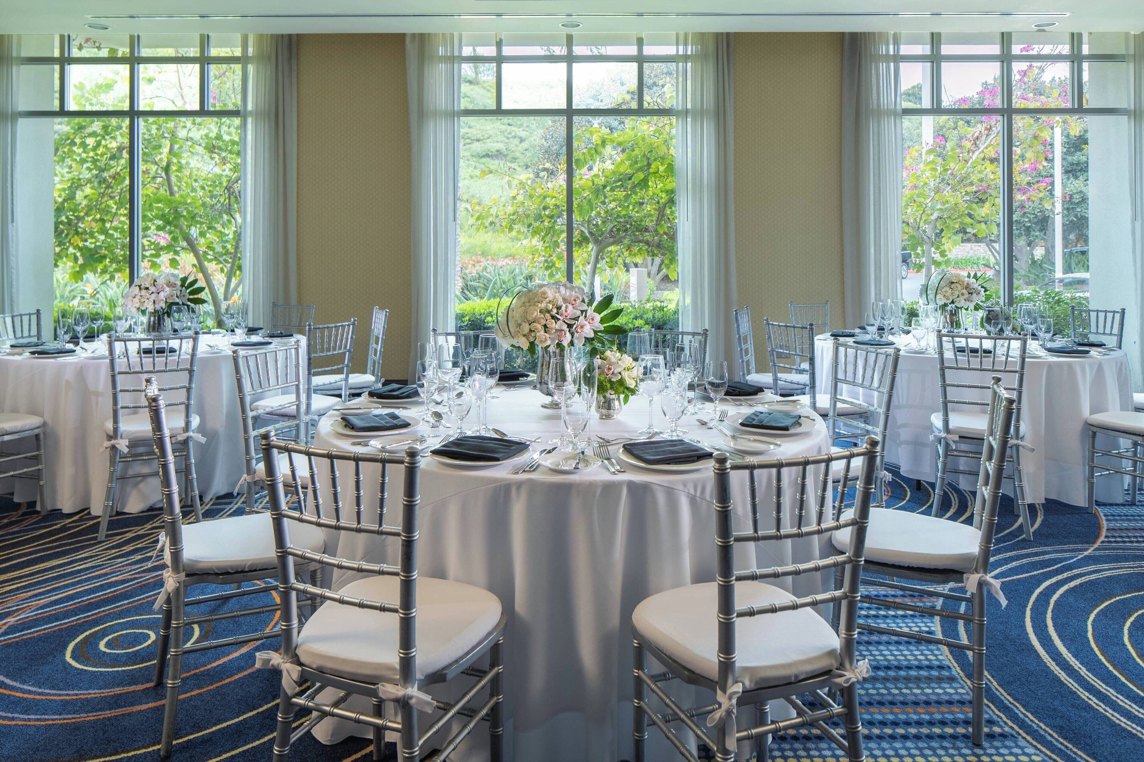 Un salón de banquetes con mesas redondas dispuestas para un evento formal, rodeado de sillas con asientos acolchados.