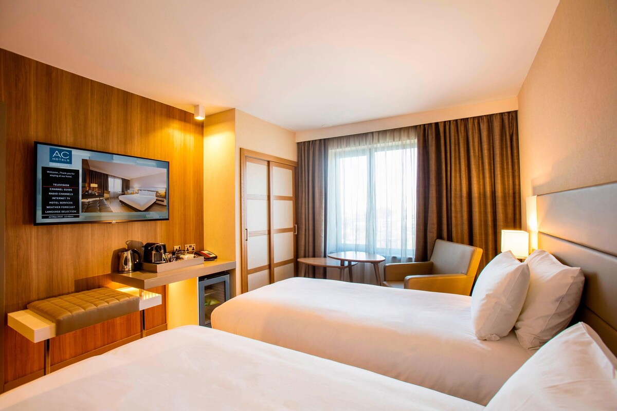Habitación de hotel con dos camas, televisión, escritorio con cafetera, una silla y ventana con cortinas.