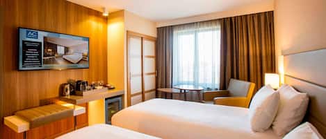 A hotel room with two beds, a TV, a desk with a coffee maker, a chair, and a window with curtains.