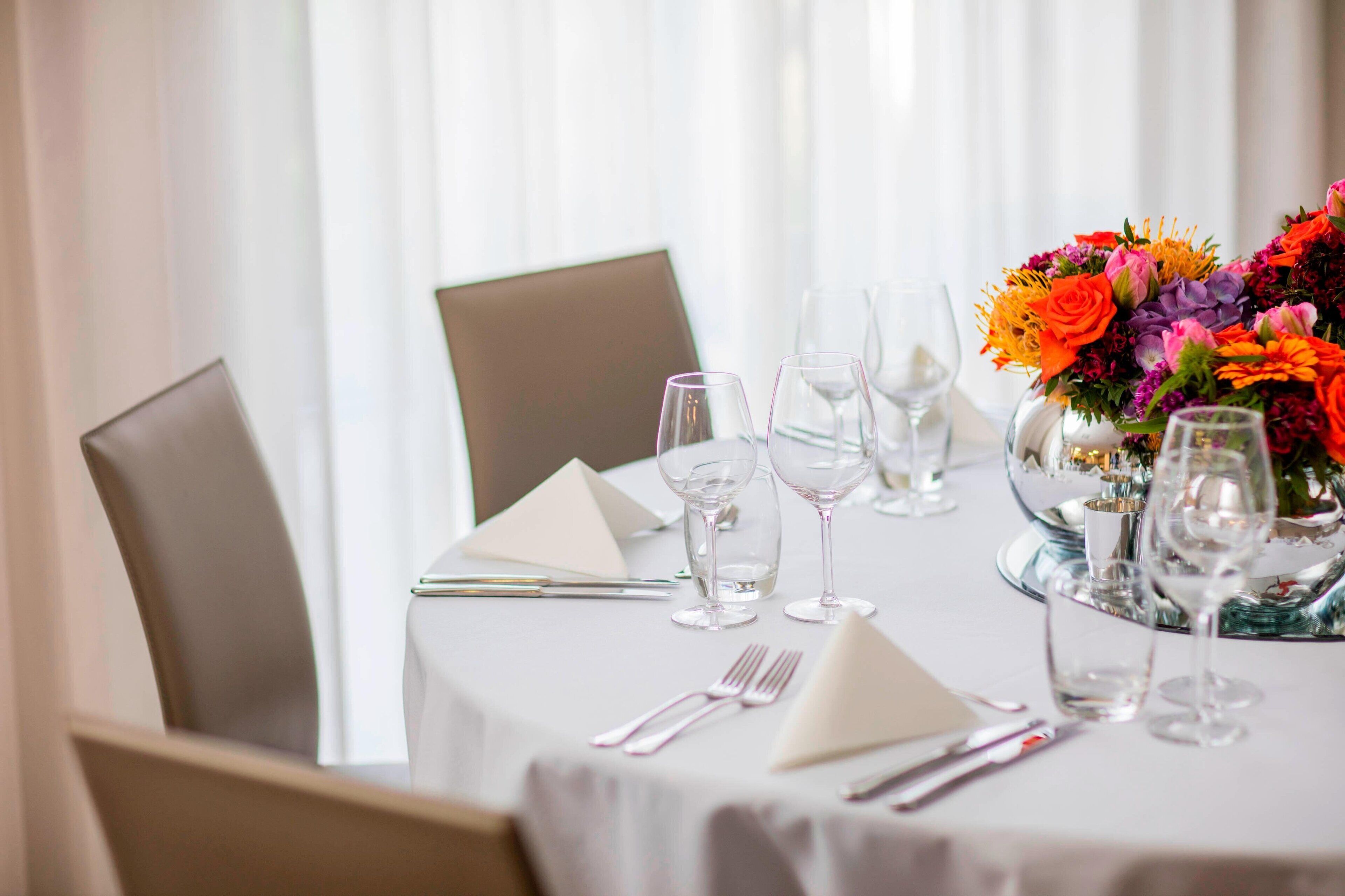 Una mesa lista para comer, con un centro de mesa floral, cristalería transparente y mantel blanco.