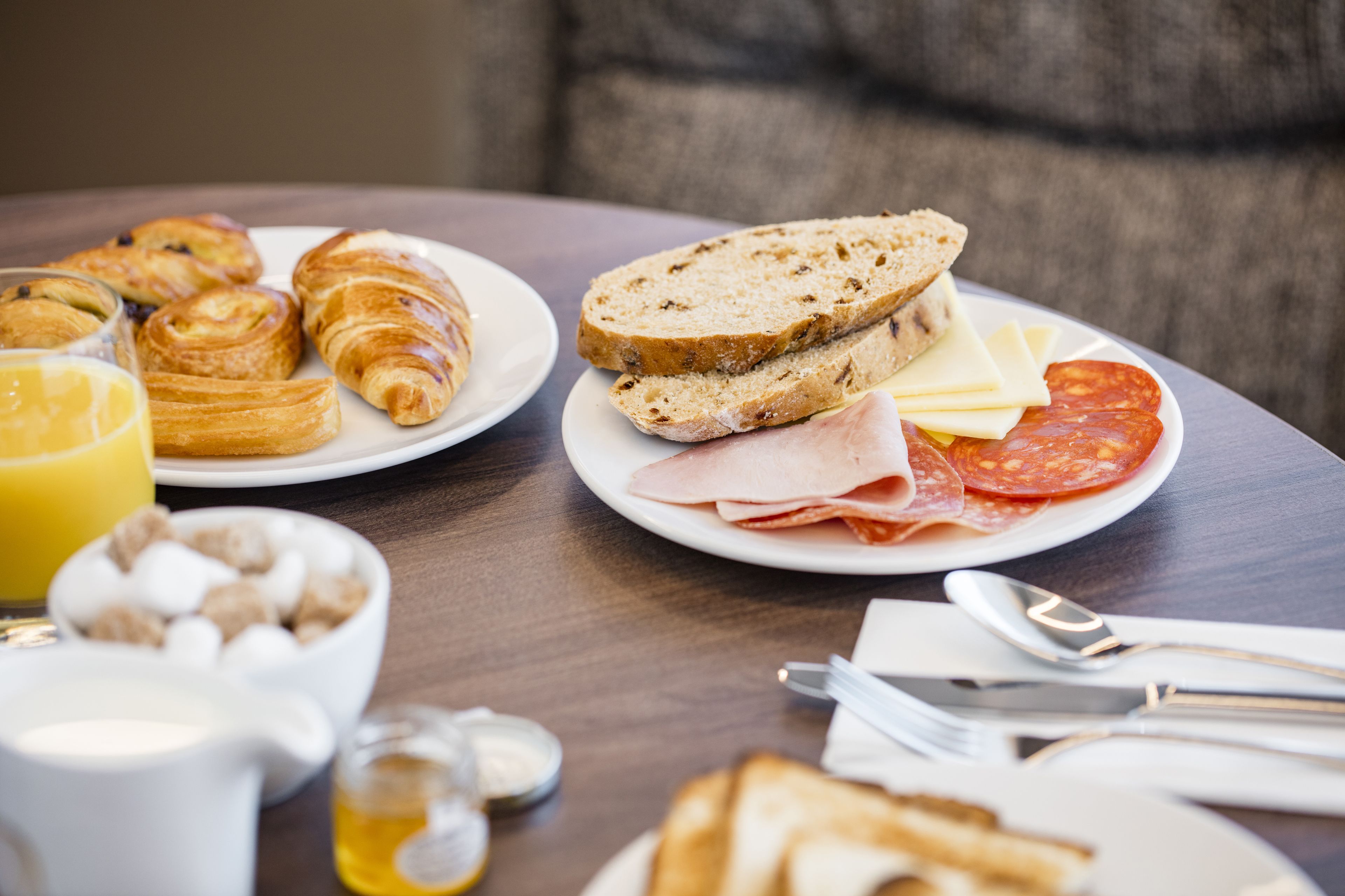 Ein Frühstückstisch mit Croissants, Brot, Käse, Schinken und Würstchen auf Tellern, dazu eine Schüssel Müsli und ein Glas Orangensaft.