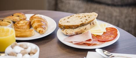 Ein Frühstückstisch mit Croissants, Brot, Käse, Schinken und Würstchen auf Tellern, dazu eine Schüssel Müsli und ein Glas Orangensaft.