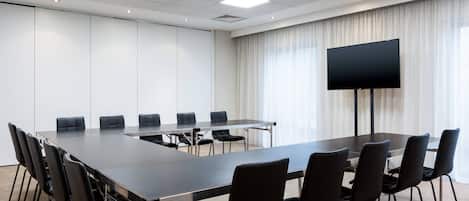 A conference room with a long table, black chairs, a large flat-screen TV, and a window with curtains.