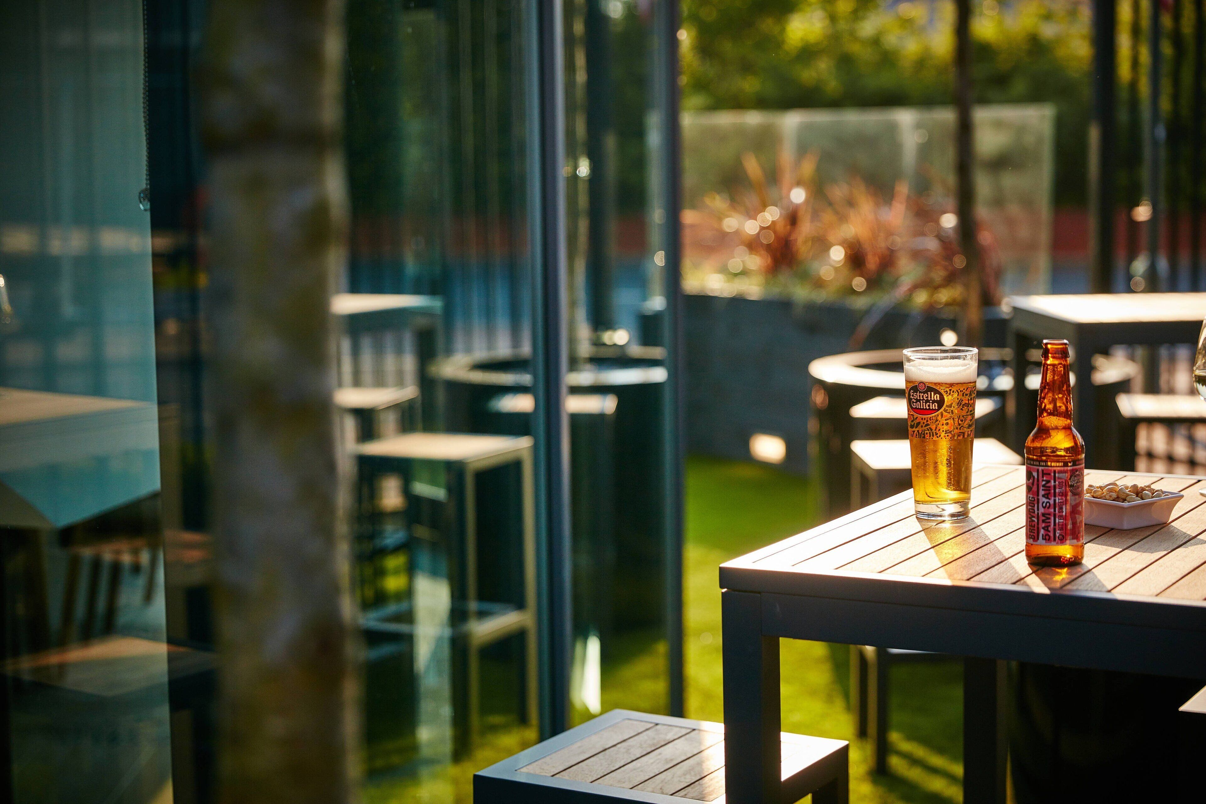 An outdoor seating area with tables and chairs, a glass partition, and a bottle of beer on one table.